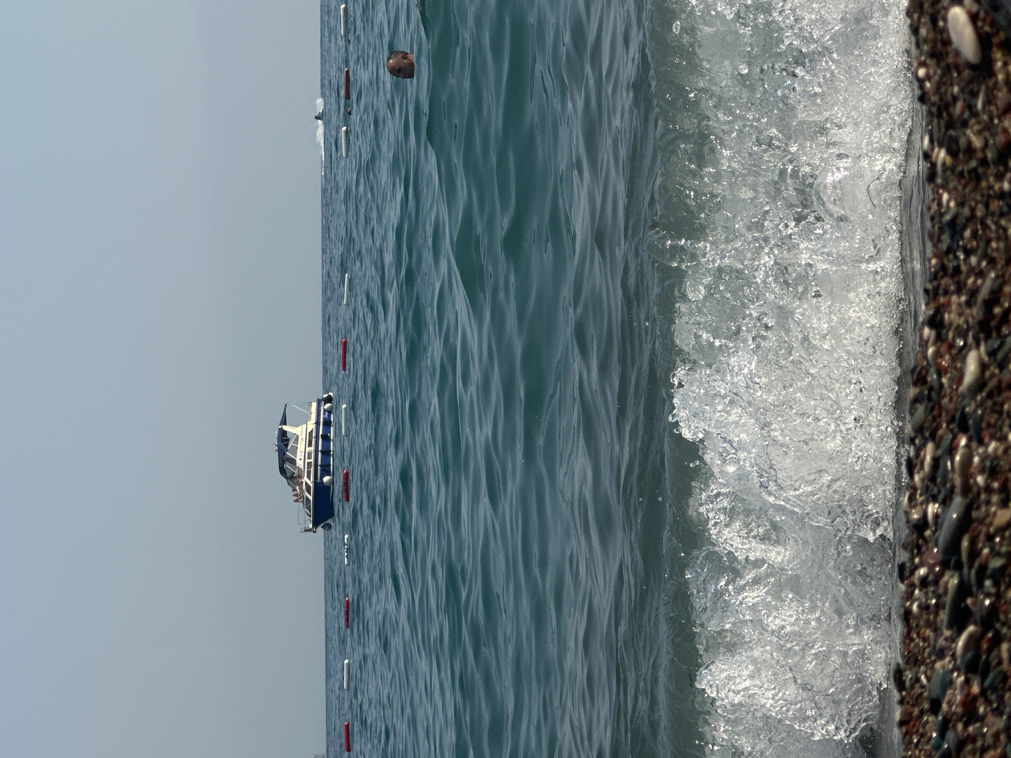 A medium shot captures a sunny day at a beach in Boreti, Montenegro. The foreground is dominated by a pebbled shoreline leading to the water's edge, where a breaking wave creates a foamy cascade. In the mid-ground, the turquoise sea stretches out, marked by a line of buoys. A man's head is visible above the water, near the buoys, appearing to be swimming or enjoying the sea. Further in the background, a white and blue motorboat with people on deck is anchored in the water. To the right of the motorboat, a person on a jet ski is visible, creating a spray of water. The sky is clear and a pale blue, indicating fair weather. The overall scene depicts a tranquil beach environment with recreational activities taking place on the water.