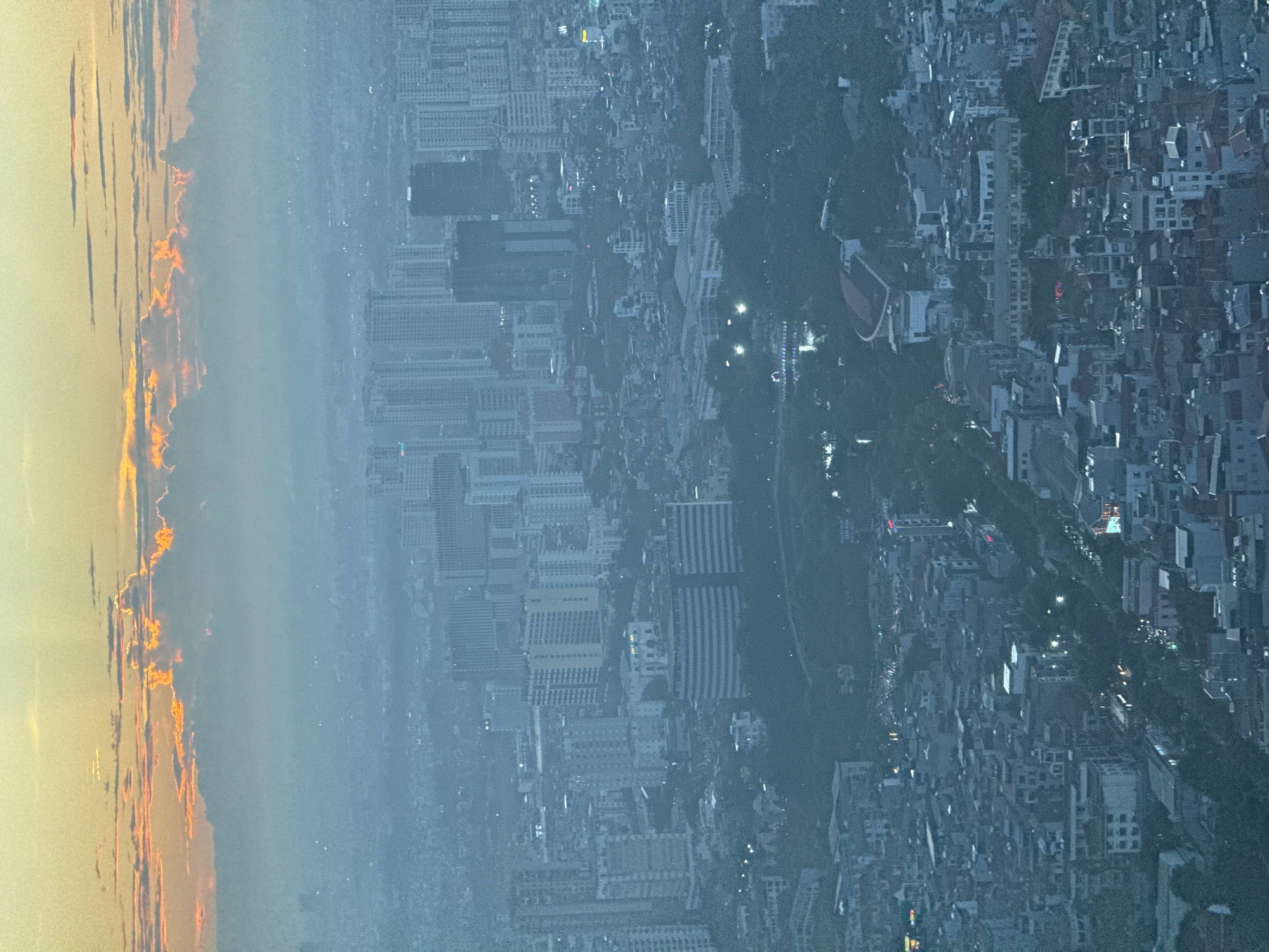 The image is an aerial view of Hanoi, Vietnam, likely captured at dusk or dawn. The sky is illuminated with warm tones of orange and yellow, with scattered clouds. The city below is shrouded in a blue-grey haze, typical of urban environments. A dense cluster of buildings, ranging in height from low-rise structures to taller, modern high-rises, fills the frame. Vegetation, in the form of trees and parks, is interspersed throughout the urban landscape. The lighting suggests that artificial lights from the city are beginning to illuminate, with some points of light visible in the distance. There are no visible people or animals in the image, but the presence of infrastructure indicates human habitation and activity. The overall impression is one of a vast, sprawling urban area under a dramatic sky.