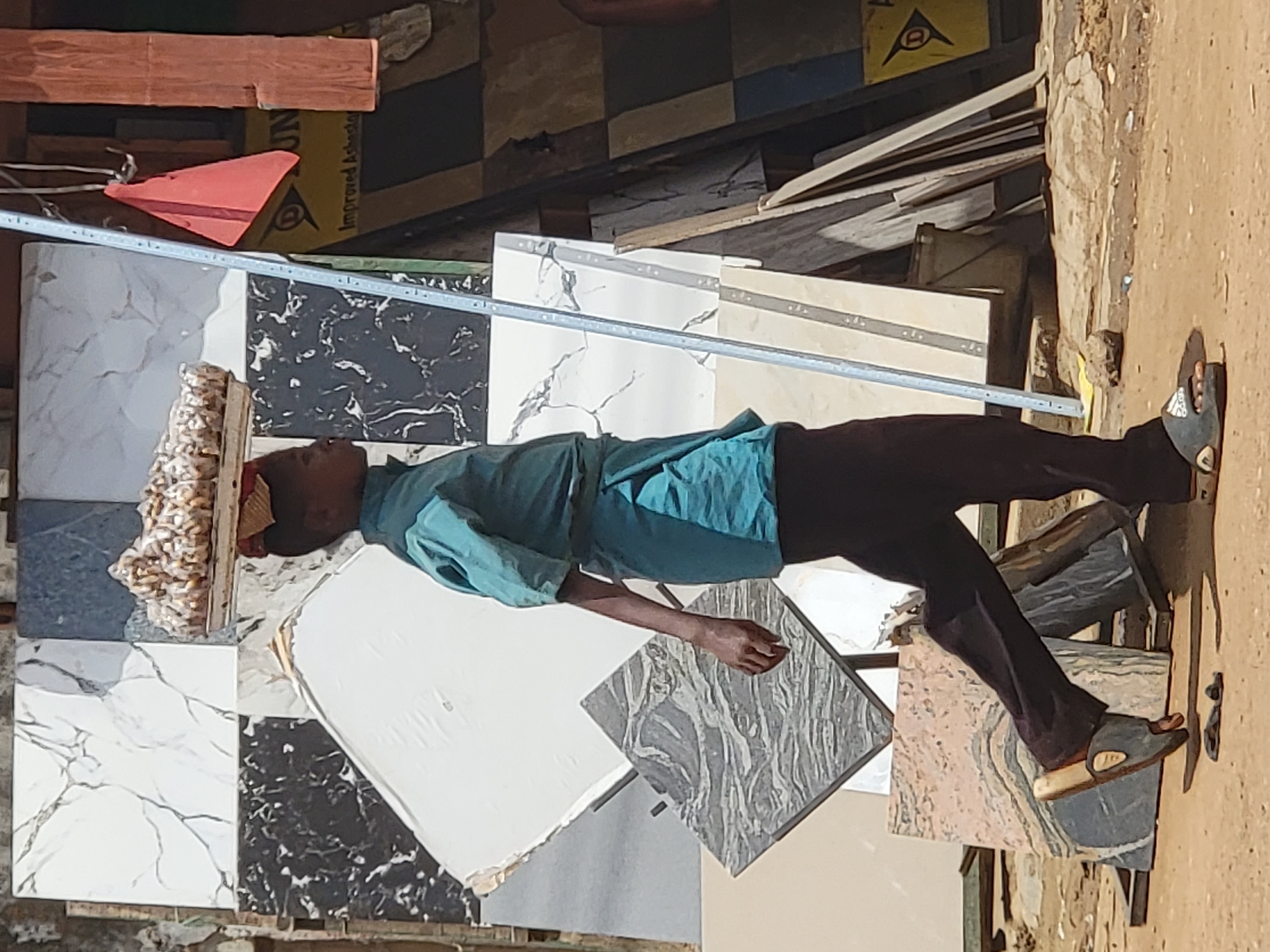 The image depicts an outdoor scene in Maiburiji, Nigeria, characterized by bright daylight. A young person, dressed in a blue-teal short-sleeved shirt, dark trousers, and sandals, is walking while expertly balancing a large, lumpy, light-colored load on their head. The person's face is partially obscured, but they appear focused on carrying their burden.
The immediate surroundings are dominated by a display or storage of various tiles, suggesting a tile shop, market stall, or construction material yard. These tiles come in an array of patterns and colors, including white with black veining, black with white veining, mottled grey, and a pinkish-beige marble-like design. Some tiles have visible silver-colored edge trims.
In the background, there are stacks of wooden planks and other construction debris. A yellow object or sign is partially visible, bearing the text "Improved Asbestos" and "DUNLOP," along with a triangular logo. The ground is unpaved, consisting of dirt or sand. The overall impression is one of a bustling commercial or work area under a clear, sunny sky.