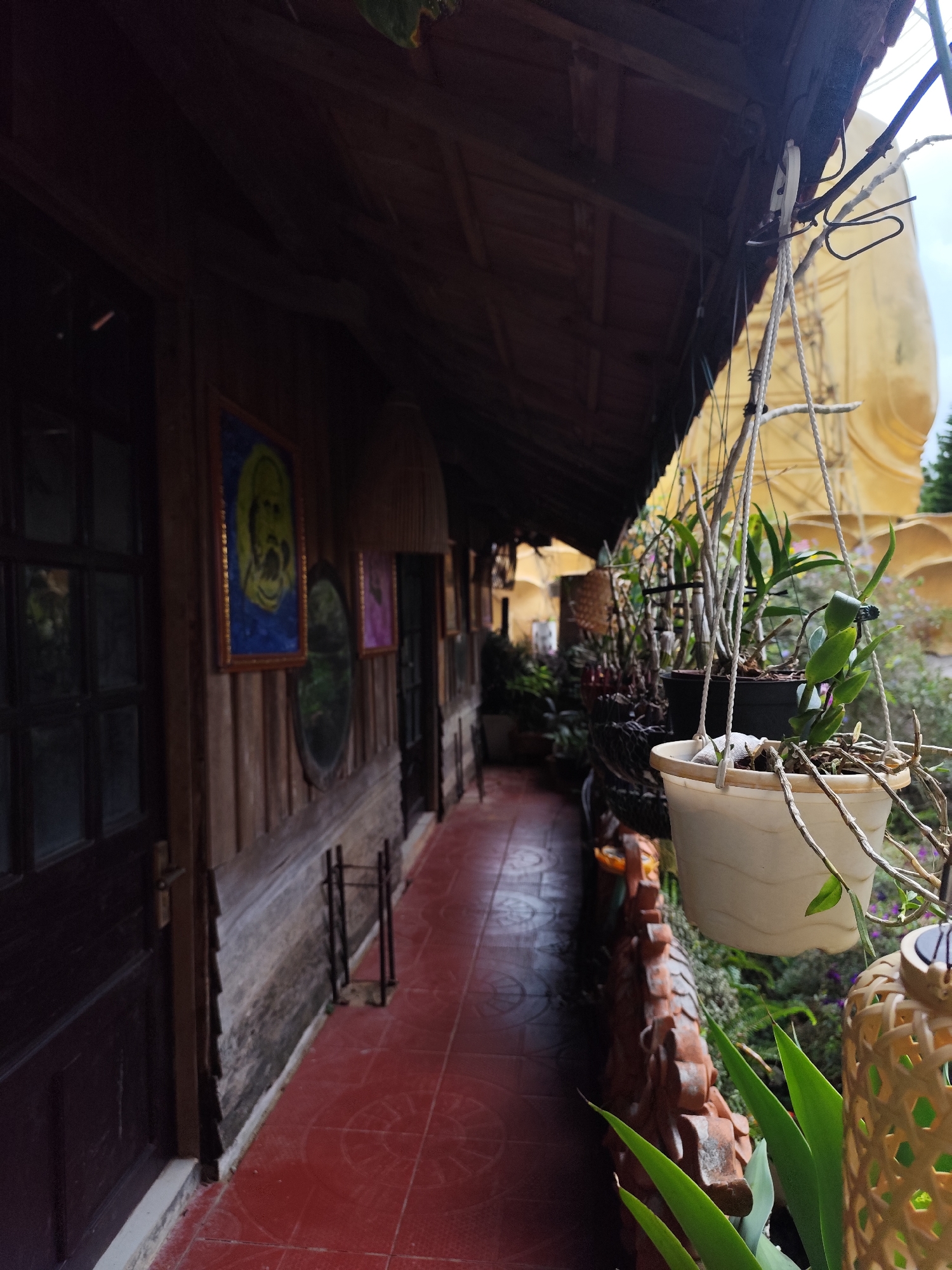 A perspective view depicts an exterior corridor in Lâm Viên - Đà Lạt, Vietnam, with a red-tiled floor. A wooden wall adorned with framed pictures and an oval mirror runs along the left. Hanging plants in pots are suspended from the wooden roof, partially obscuring a large, golden Buddha statue in the background. Additional foliage is visible below the railing. The lighting is overcast.