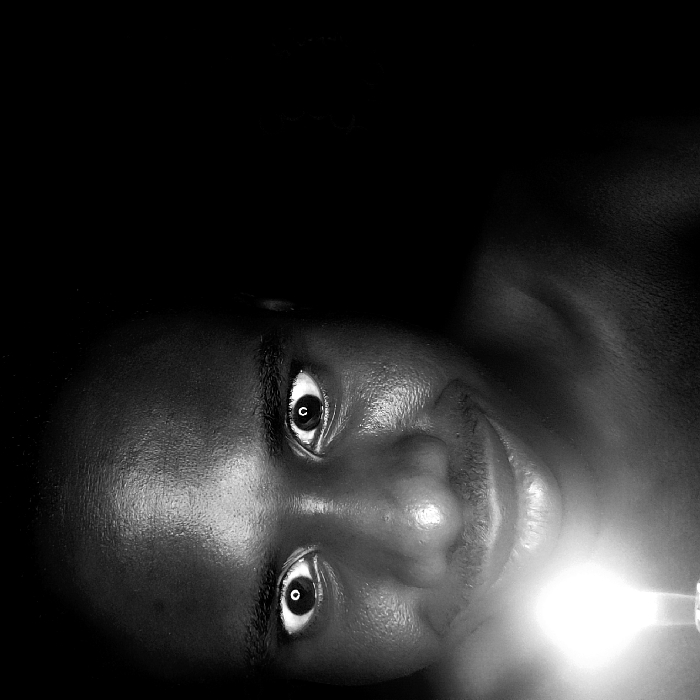 A black and white close-up image depicts a male individual, appearing to be of African descent, located in Narayi, Nigeria. The subject's face is illuminated from below by a single, bright light source, consistent with a lit candle, held out of frame below his chin. His eyes are wide open, pupils visible, and he exhibits a slight smile or smirk. Strong highlights are present on his forehead, nose, and cheekbones due to the upward light. The background is completely dark, suggesting an indoor setting at night or in a room devoid of other illumination. The individual appears bare-chested or wears dark attire that blends with the background.