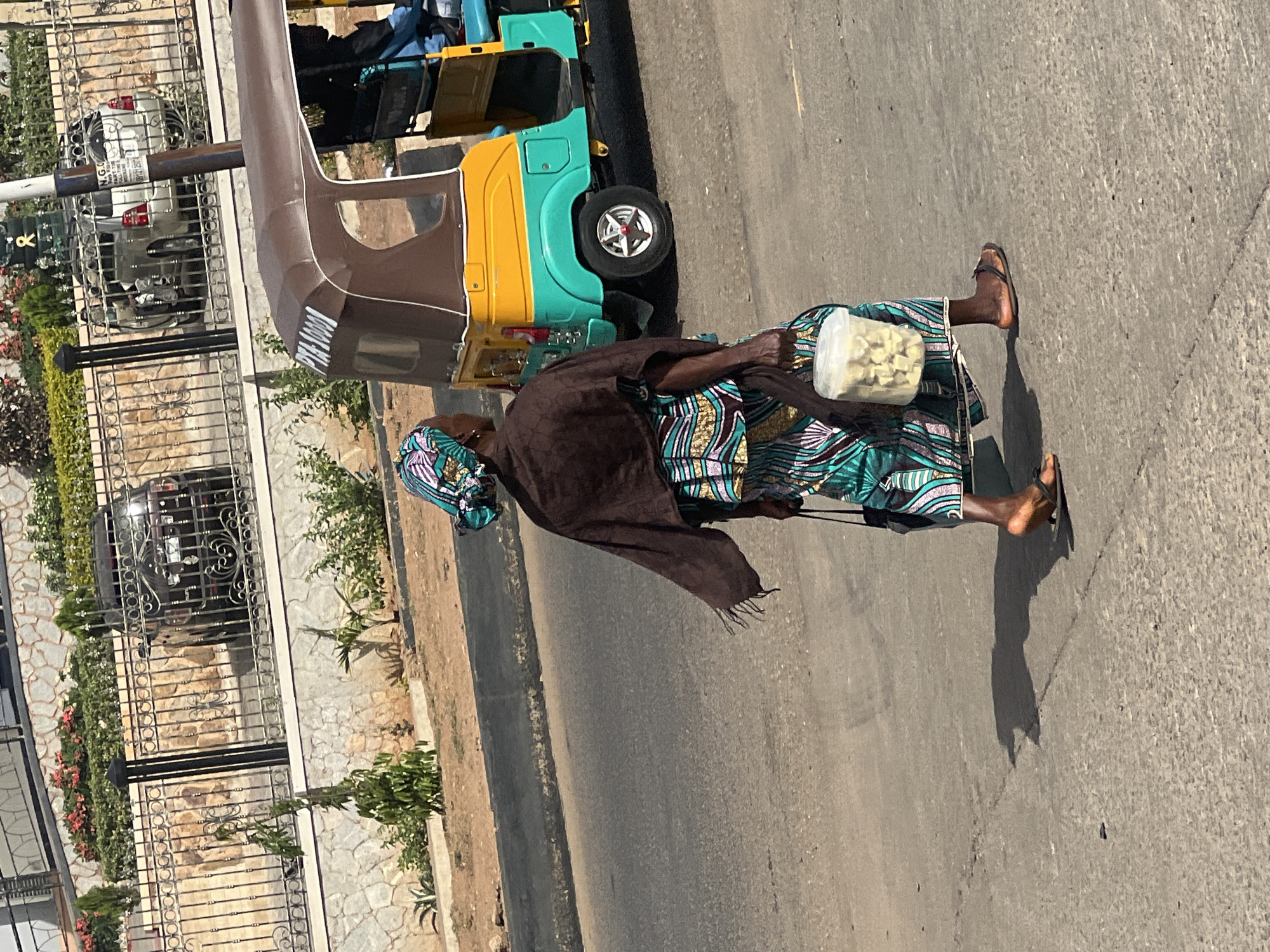 The image depicts a street scene in what appears to be Jos, Nigeria, under bright, clear daylight conditions, indicated by the strong shadows cast on the asphalt road. The primary subject is an adult woman walking from right to left across the frame. She is dressed in traditional attire, including a vibrant, patterned long dress or wrapper in shades of teal, gold, and white, a matching headscarf, and a dark brown shawl draped over her shoulders. She is wearing dark flip-flops and is carrying a clear plastic container filled with small, light-colored, cuboid food items, suggesting she might be a street vendor.
To the woman's right, a colorful motor tricycle, commonly known as a "keke napep," is prominently featured. It has a brown canopy over the passenger section, with a yellow and teal body. The word "WELCOME" is visible in white letters on the brown part of the canopy. The tricycle appears to be either parked or moving alongside the woman.
In the background, behind a black ornate metal fence, there are several parked cars, including a silver sedan. On the silver car, or a sign near it, the text "N.G.S" and partial phone numbers, including "070 125", are visible. Beyond the fence, there is a paved area with stone patterns and various green plants and bushes, suggesting a residential or commercial property. The scene captures an everyday moment of urban life.