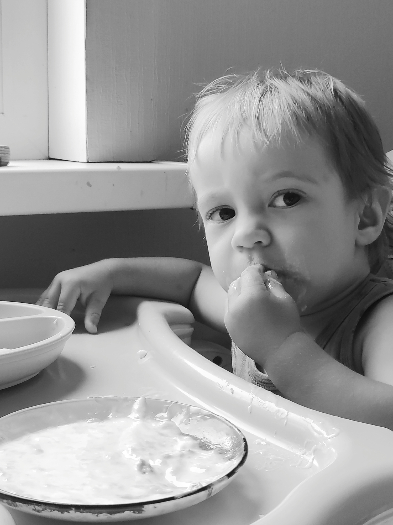 In Chaikovsky, Russia, a young child, estimated to be around 1-2 years old, sits in a high chair, engaged in eating. The child has light-colored hair and is looking directly at the camera with wide, dark eyes. Their right hand is raised to their mouth, with fingers inside, suggesting they are self-feeding or exploring the food. The child's face and hand are smudged with food.
In front of the child is a white high chair tray. On the tray, there are two white bowls, one partially visible on the left and another in the foreground containing a white, creamy substance that appears to be food. The tray itself has some food residue. The background consists of a wall and a window frame, with a windowsill. The lighting suggests an indoor setting, likely a home environment. The overall image is captured in black and white, emphasizing textures and light.