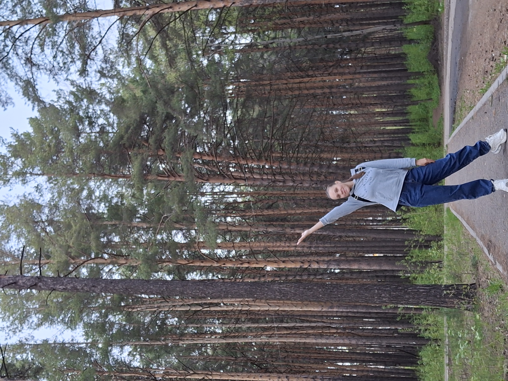 The image shows a woman standing on a pathway in a pine forest. She is wearing jeans, a gray sweatshirt, and white sneakers. Her left arm is extended outward. The pathway is made of concrete and runs alongside a grassy area. The trees are tall and dense, with thick brown trunks and green needles. The location is the city of Chaikovsky, Russia.