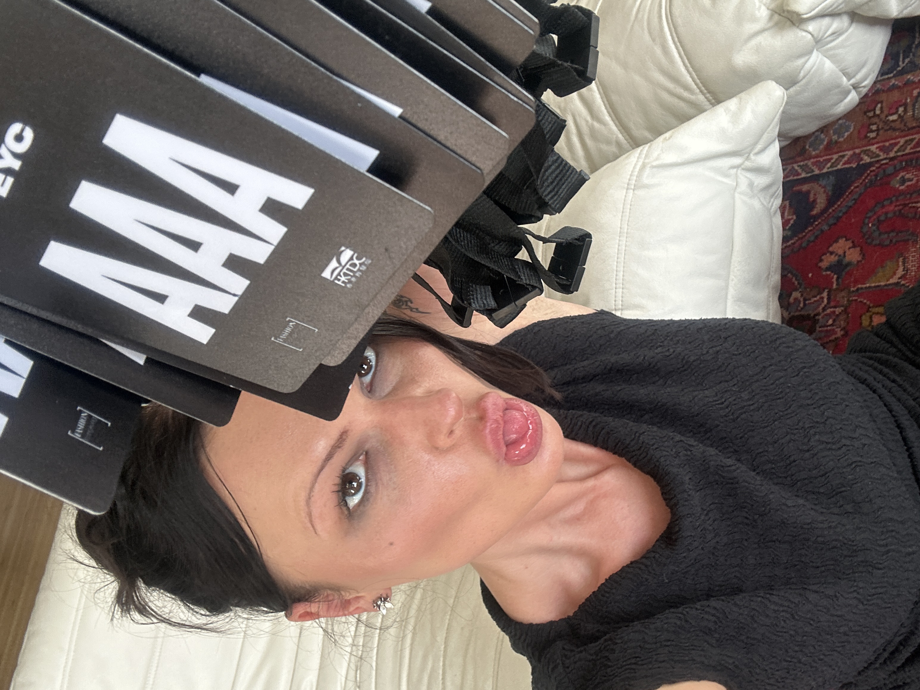 A close-up, vertical photograph captures a female subject from the chest up within an indoor setting. The woman has dark hair styled in a messy bun and wears a black, textured, short-sleeved top. She looks directly into the camera with brown eyes and a playful expression, her tongue slightly extended. A small, metallic, floral or insect-shaped earring is visible on her right ear, and a partial tribal-style tattoo is visible on the right side of her neck.
She holds a stack of black identification badges attached to lanyards in the upper right portion of the frame. The badges feature prominent white text "AAA", and smaller visible text includes "FASHION" within brackets and the logo "HKTDC" with associated Chinese characters, suggesting involvement with the Hong Kong Trade Development Council at a fashion event. The letters "EYC" are partially visible at the top of one badge.
The background primarily consists of a light-colored, quilted upholstered surface, possibly a headboard or sofa. A section of a red and blue patterned oriental rug is visible at the bottom right. The scene depicts an individual associated with a fashion event, located in the city of London, United Kingdom.