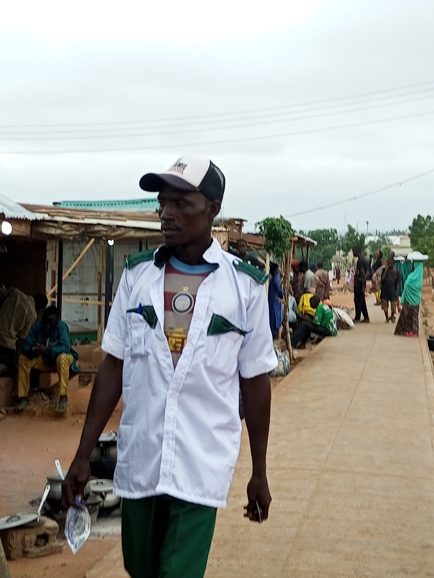 In Pantame, Nigeria, a man wearing a white short-sleeved shirt with green epaulets and a black and white baseball cap walks along a dirt pathway. He is wearing dark green trousers and a striped t-shirt underneath his open white shirt. He holds a clear plastic bag in his right hand. In the background, several other people are visible, some sitting and some standing. The environment appears to be a marketplace or a gathering area with simple structures, possibly made of corrugated metal and wood. The ground is dusty. The sky is overcast.