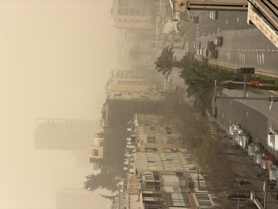 An elevated view captures an urban thoroughfare in Tel-Aviv, Israel, enveloped in a dense, uniform yellowish-brown dust storm that severely limits visibility. The entire sky is obscured by this haze, creating diffused light.
In the foreground, the lower right exhibits a concrete ledge or balcony edge, indicating the elevated photographic position. Below, a wide, multi-lane street, likely a boulevard, extends into the hazy distance, accommodating traffic in both directions. A central median strip features several mature palm trees with green fronds. Numerous vehicles are present; some are parked along the left curb, while others are in motion within the traffic lanes. A traffic light in the center-right is illuminated green. Sidewalks border the street on both sides. On the left sidewalk, several bare-branched deciduous trees and streetlights are visible, along with a single pedestrian walking away from the viewer. In the right-middle distance, a construction site with excavated earth and debris is discernible through the dust.
Multi-story buildings line both sides of the street. On the left, buildings vary in style and color, mostly light-toned (white, beige, pale yellow), many featuring balconies. One prominent building in the mid-left appears to be under construction or renovation, with dark netting or scaffolding covering a significant portion of its facade. A dark green tree, possibly coniferous, stands behind these buildings. In the background, visibility diminishes further, with faint silhouettes of numerous multi-story buildings. A very tall, slender, partially constructed high-rise building with an exposed skeletal frame is visible as a prominent silhouette in the far background, confirming a major urban development area.