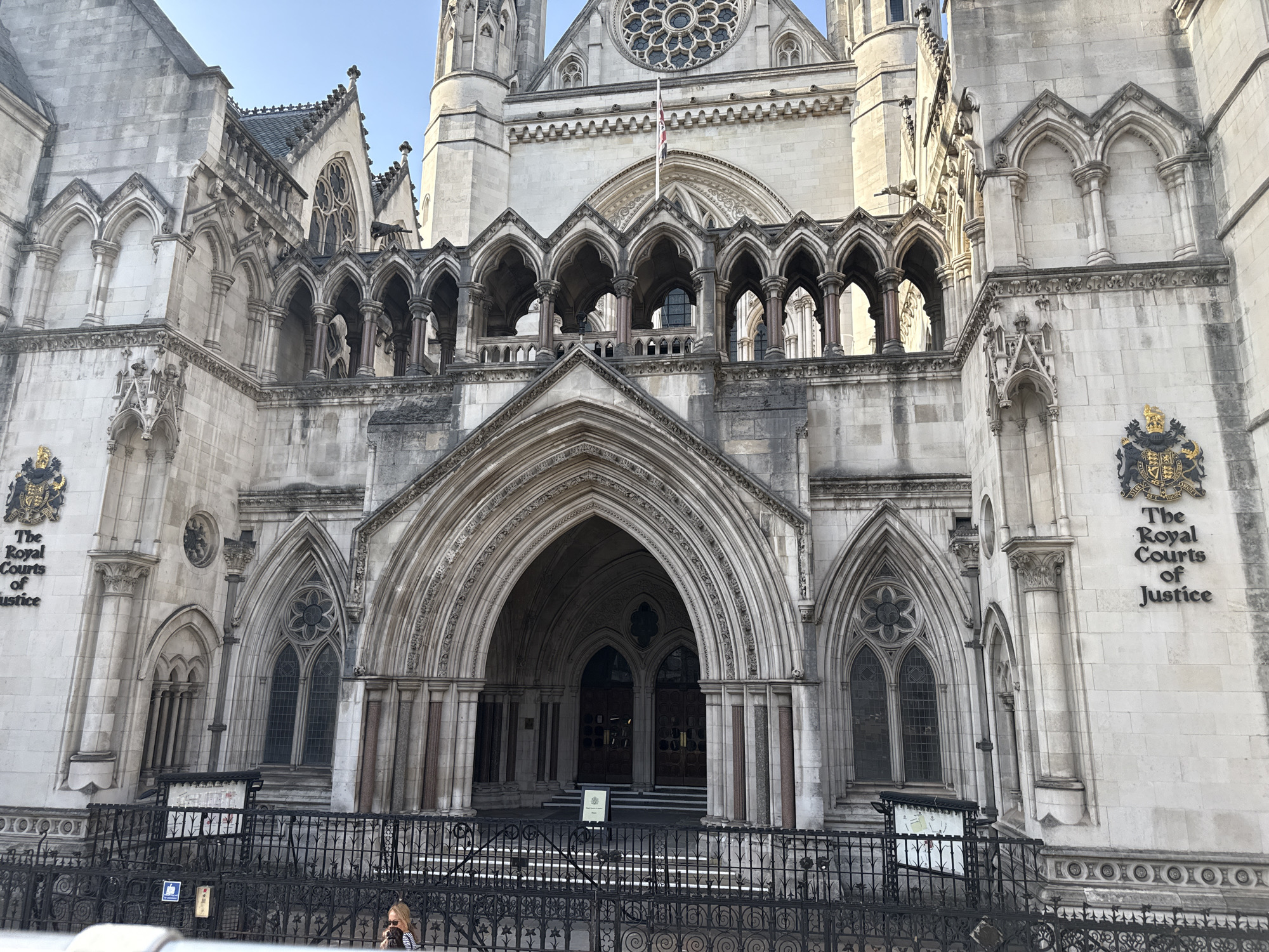 The image shows the Royal Courts of Justice in London, United Kingdom. It is a large, ornate building with Gothic architectural details. The facade features multiple arches, columns, windows, and stone carvings. The entrance is framed by a large pointed archway. A black wrought-iron fence runs in front of the building, separating it from the street. There are signs with maps. A person can be seen behind the fence on the left side of the image. The sky is blue.