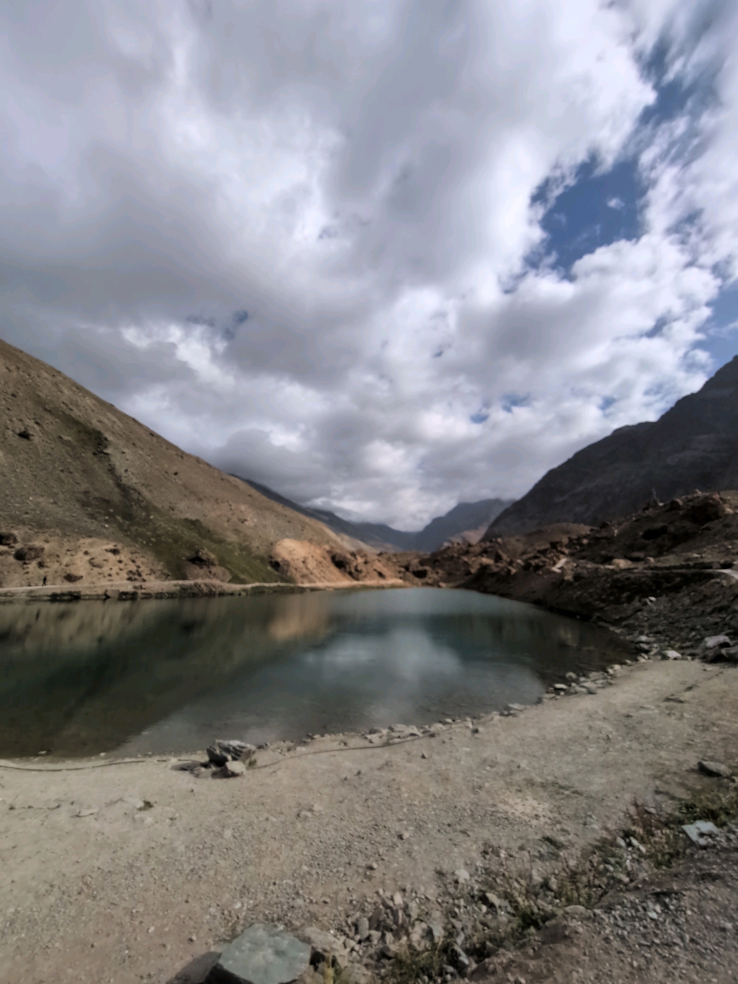 A scenic view of the Chandra Tal lake in Patseo, India, showcases the serene natural beauty of the region. The image captures the vast expanse of the lake, its calm waters reflecting the dramatic interplay of light and shadow cast by the overcast sky. Surrounding the lake are steep, barren mountains, their rugged slopes a testament to the harsh yet beautiful terrain. The foreground is characterized by a sandy, rocky shoreline, with scattered stones and patches of dry grass indicating a remote, arid environment. In the distance, layered mountains fade into the horizon, creating a sense of depth and vastness. The sky is a dynamic canvas of clouds, with breaks of blue sky peeking through, hinting at the changing weather conditions typical of high-altitude regions. The overall atmosphere is one of tranquility and raw, untamed nature, drawing the viewer into the peaceful solitude of this Indian mountain landscape.