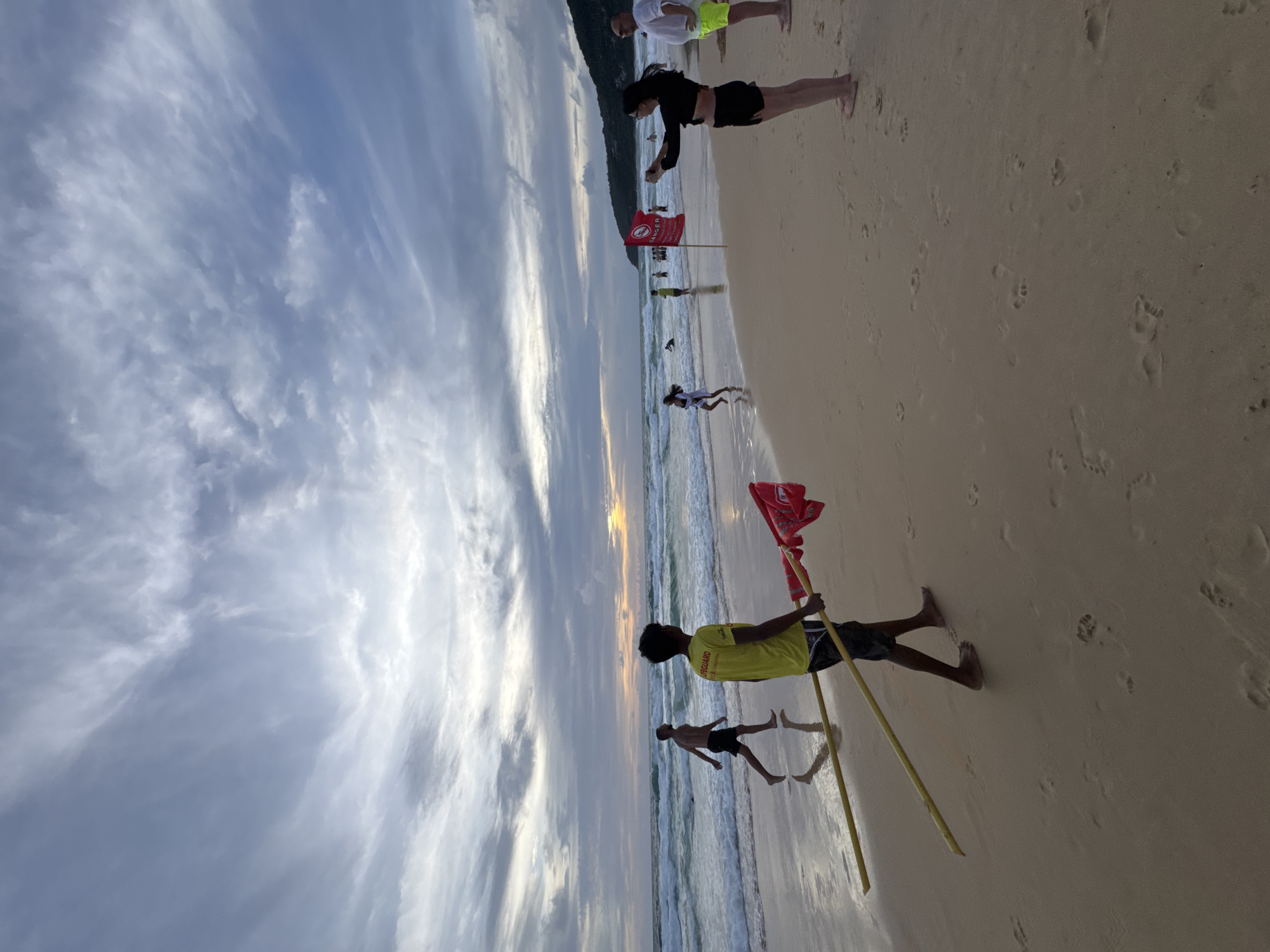 A lifeguard in a yellow shirt carries two yellow poles with a red flag attached to them on a beach in Karon, Thailand. The lifeguard walks towards the right side of the image. The beach has wet sand with footprints. In the background, waves crash on the shore, and the ocean stretches to the horizon. Several people are visible on the beach and in the water, some walking and others swimming. A red "DANGER" flag is planted in the sand on the right side of the image. The sky is cloudy with hints of sunset colors visible.