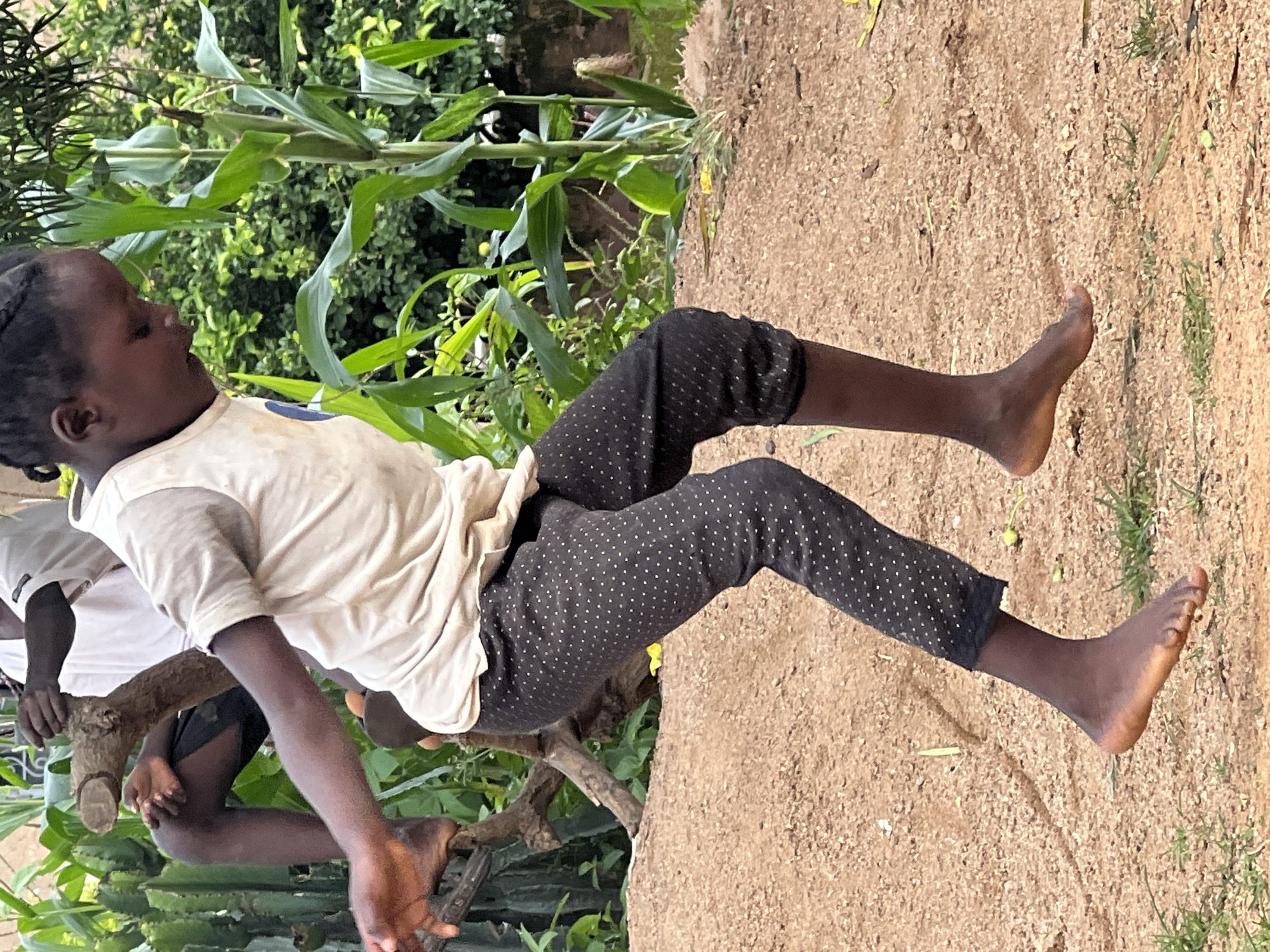 The image shows a young Black child in a light-colored t-shirt and dark patterned pants, sitting outdoors. The child is on the left side of the frame, with their body angled towards the right. Their right leg is extended, and their right foot is visible, with toes spread. The child's left leg is bent, and their left foot is also visible, tucked beneath their body. The child has braided hair.
Behind the child, the scene consists of a dirt ground surface with some sparse green grass. In the background, there are tall green plants, possibly corn stalks, and other foliage. A tree branch is visible in the foreground, with the child’s arm resting on it. Another child or person is partially visible to the left, wearing a white shirt.
The lighting suggests it is daytime, likely with sunlight casting shadows. The overall setting appears to be a rural or natural environment. The child’s expression is not clearly visible, but their body posture might suggest a moment of rest or play. There is no visible text in the image. The location context is Jos, Nigeria.