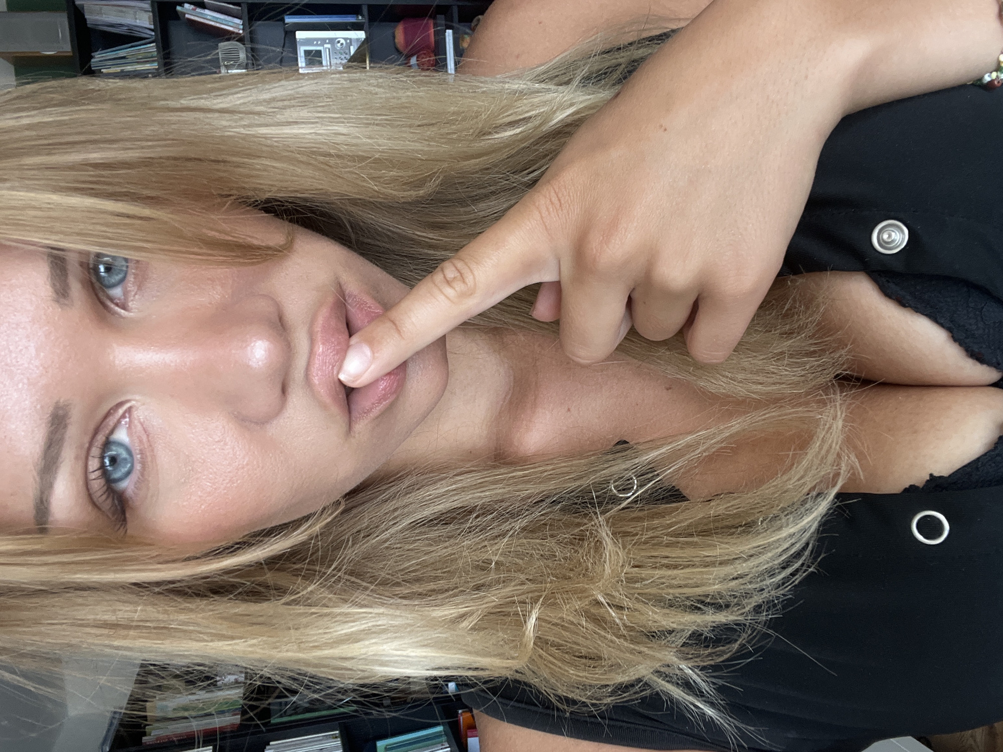 A close-up, horizontally oriented, over-the-shoulder shot shows the face of a young woman with blonde hair and blue eyes. She is touching her lips with her right index finger, and her gaze is directed towards the camera. Her lips are slightly parted and appear to be painted with a light pink lipstick. Her skin has a subtle glow. She is wearing a black top with metallic eyelets. The background is slightly out of focus and shows bookshelves filled with books and other items. The lighting suggests an indoor setting, possibly during daylight hours. The overall mood is intimate and engaging. No visible text is present in the image.