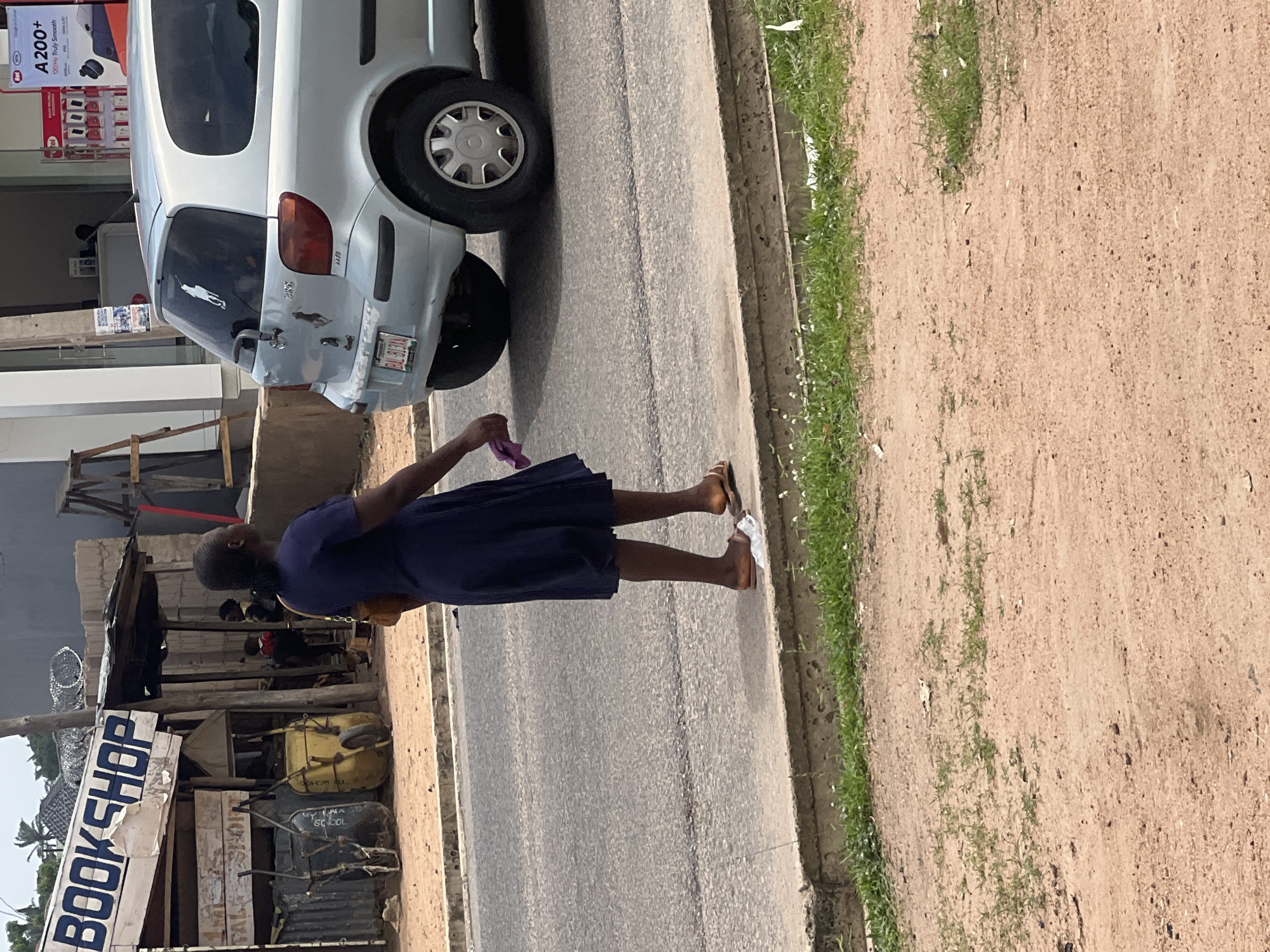 The image depicts a street scene in Jalingo, Nigeria, on a slightly overcast day. In the foreground, a woman wearing a dark blue dress and brown sandals stands on the side of a paved road, holding a pink cloth in her right hand. She is facing away from the camera. To her left, a light-colored car, possibly a Toyota Vitz, is parked. The car has stickers on its rear window and a license plate that appears to read "BAL 813AA."
Behind the woman and the car, there is a modest roadside structure that appears to be a small shop. The shop has a sign with blue lettering that reads "BOOKSHOP." The structure is made of wood and corrugated metal, with items like wheelbarrows displayed in front. Further in the background, a building with a white facade and darker elements is visible, along with some vegetation and a ladder leaning against the building. The ground on the side of the road is a mix of dirt and sparse grass.