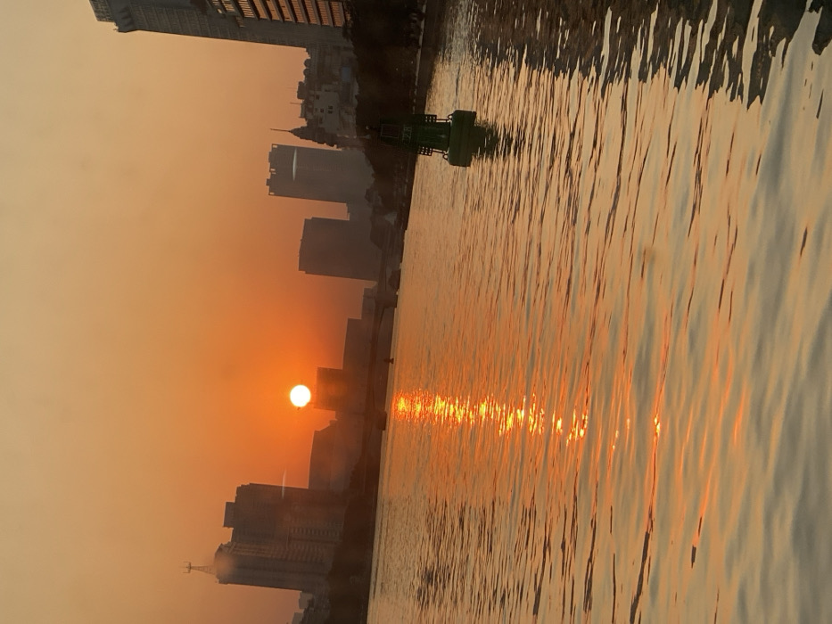The image depicts a serene sunset or sunrise over a city, likely Guang Zhou Shi, China, with a prominent body of water in the foreground. The sky is bathed in warm, intense orange and yellow hues, indicative of the golden hour.
The sun, a bright white orb, is positioned low on the horizon, partially obscured by the silhouettes of several urban buildings. Its powerful light casts a warm, hazy glow across the entire atmosphere. The city skyline, consisting of numerous high-rise buildings and structures, stands against the bright sky, appearing as dark, misty shapes on the left side of the frame.
The right side of the image is dominated by the calm surface of the water, which beautifully reflects the sun's brilliant light. A shimmering, vertical column of bright orange and yellow light extends across the water from the sun, creating a striking focal point. The water's surface shows subtle ripples and textures, enhancing the reflective patterns.
In the middle ground, a distinctive green buoy or navigational marker floats on the water, providing a contrast to the warm color palette of the scene. The overall impression is one of peaceful urban beauty, with natural light interacting dramatically with the city and water. There is no visible text in the image.