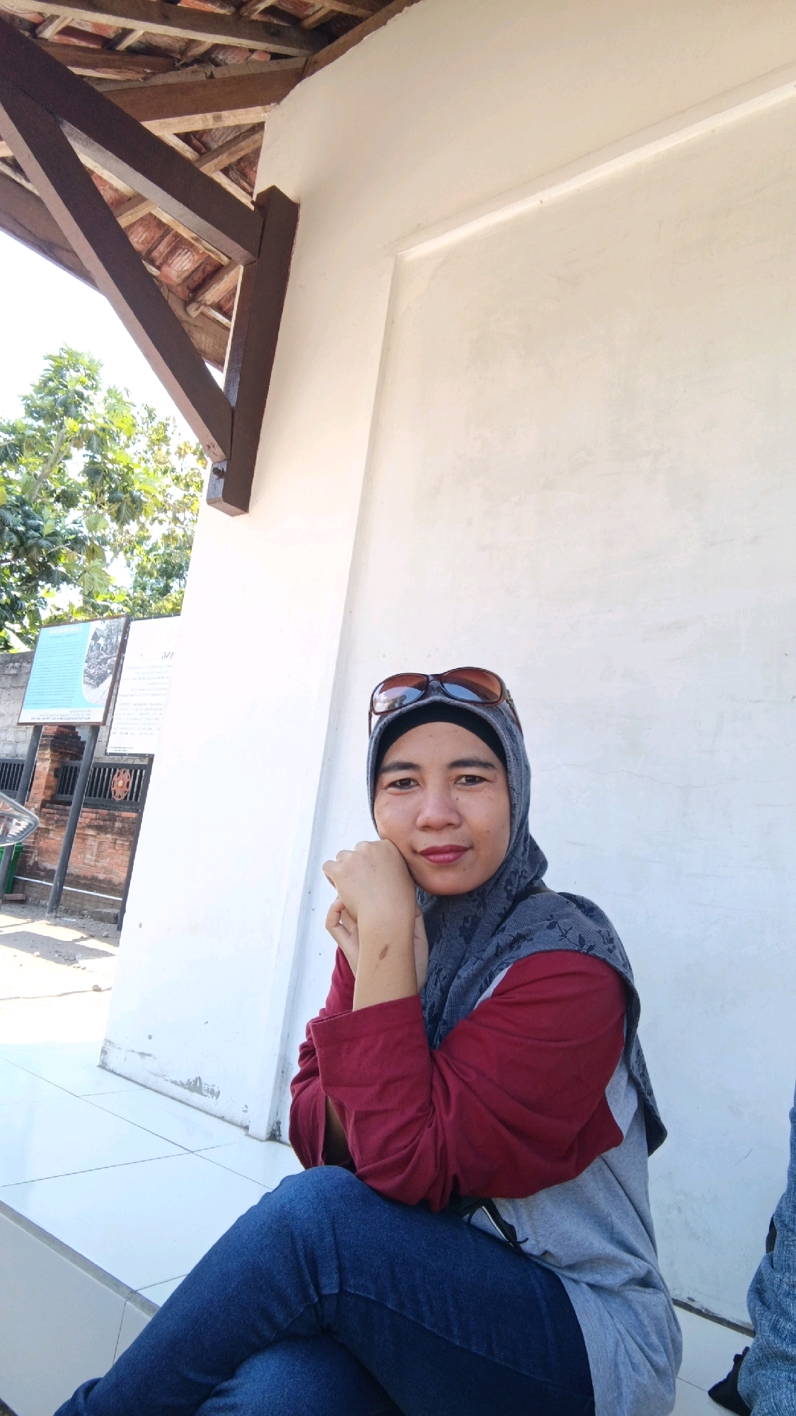 A woman, likely Indonesian, is seated outdoors against a white wall. She wears a grey hijab, sunglasses perched on top, a maroon long-sleeved shirt over a grey shirt, and blue jeans. Her hands are clasped under her chin, and she is looking towards the camera with a slight smile. Behind her, the roof structure of a building is visible, along with trees and what appear to be informational signs. The overall setting suggests a public area, possibly a historical site or park, given the signage and architecture. The image was taken during daylight.
