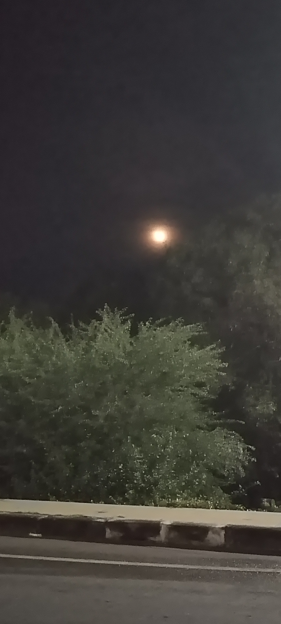 The image shows a night scene in Lhokseumawe, Indonesia. A full moon is visible in the dark sky above trees. The bottom of the image depicts a concrete curb and a section of road. There are no people visible in the image.