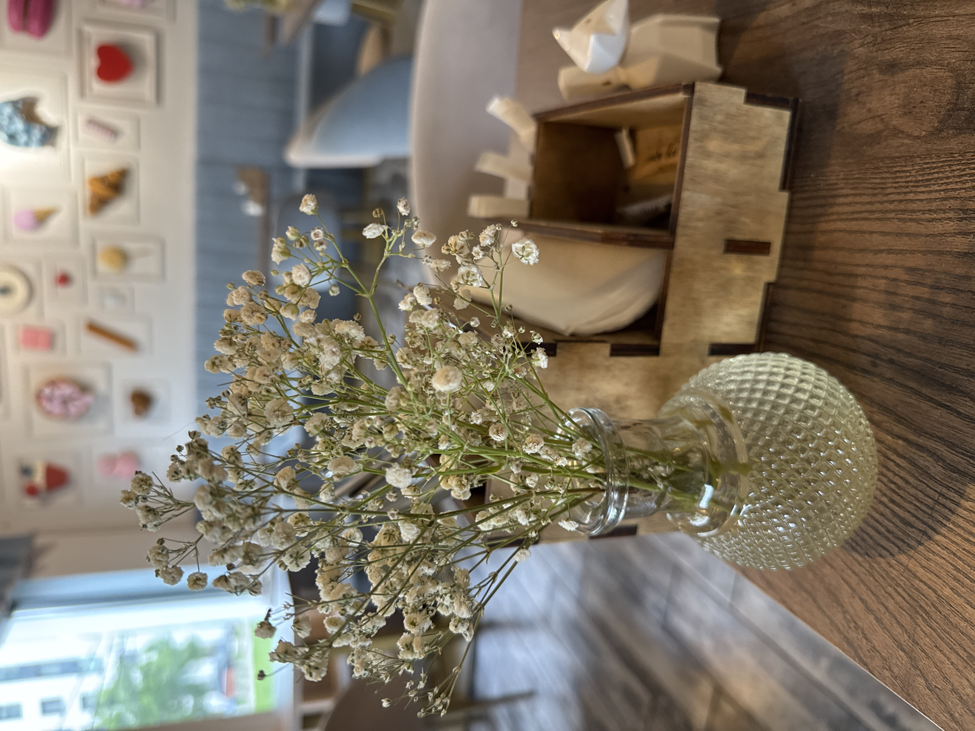 A close-up shot shows a small glass vase filled with delicate white flowers, likely Baby's Breath, sitting on a wooden table. Next to the vase is a small wooden box, containing a white folded object, possibly a napkin or small gift. The table has a dark wood grain. In the background, slightly out of focus, a wall is decorated with framed images of sweets and pastries. Through a window, greenery can be seen, suggesting an outdoor space. Given the information, the location is possibly a cafe or restaurant in Saint Petersburg, Russia.
