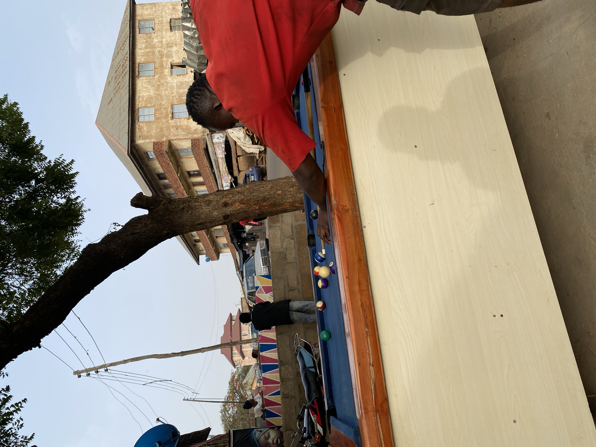 In Bukuru, Nigeria, an outdoor billiards table is the focus of activity. A man in a red shirt is bent over the table, presumably playing, while another person stands near the middle of the table observing. A motorcycle is parked nearby. The background includes a multi-story building, a tree, and utility lines.