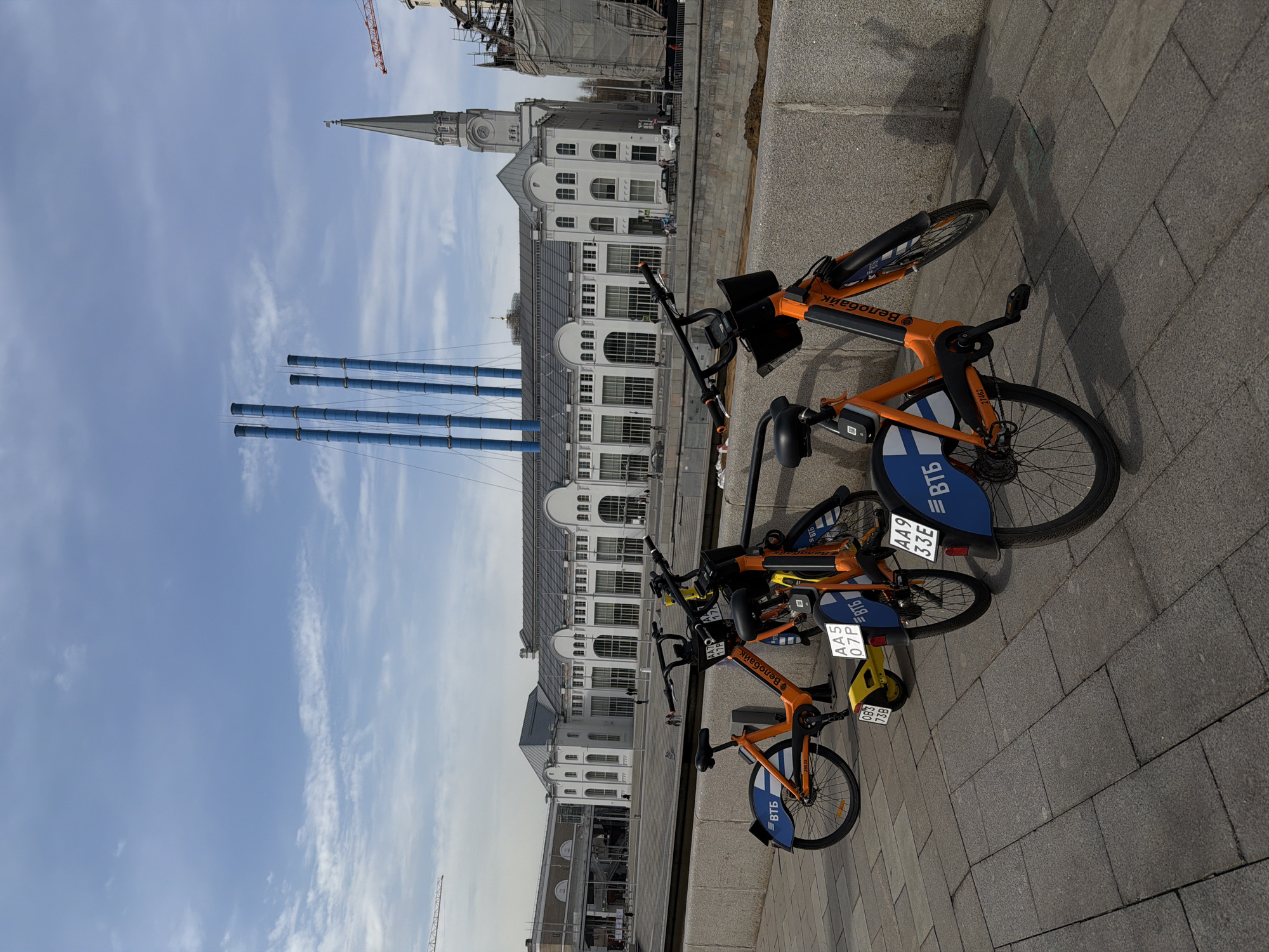 This image shows three orange and blue bicycles parked on a gray paved area. The bicycles are "Velobike" rental bikes, a bike sharing program in Moscow, Russia, as indicated by the inscriptions on the bikes. Behind the bicycles, there is a large white building with a gray roof and many arched windows. Further in the background, there are blue chimneys and a building with a tall spire. The sky above is blue with some white clouds. There is also a construction crane visible in the background. The photograph appears to be taken during the day.