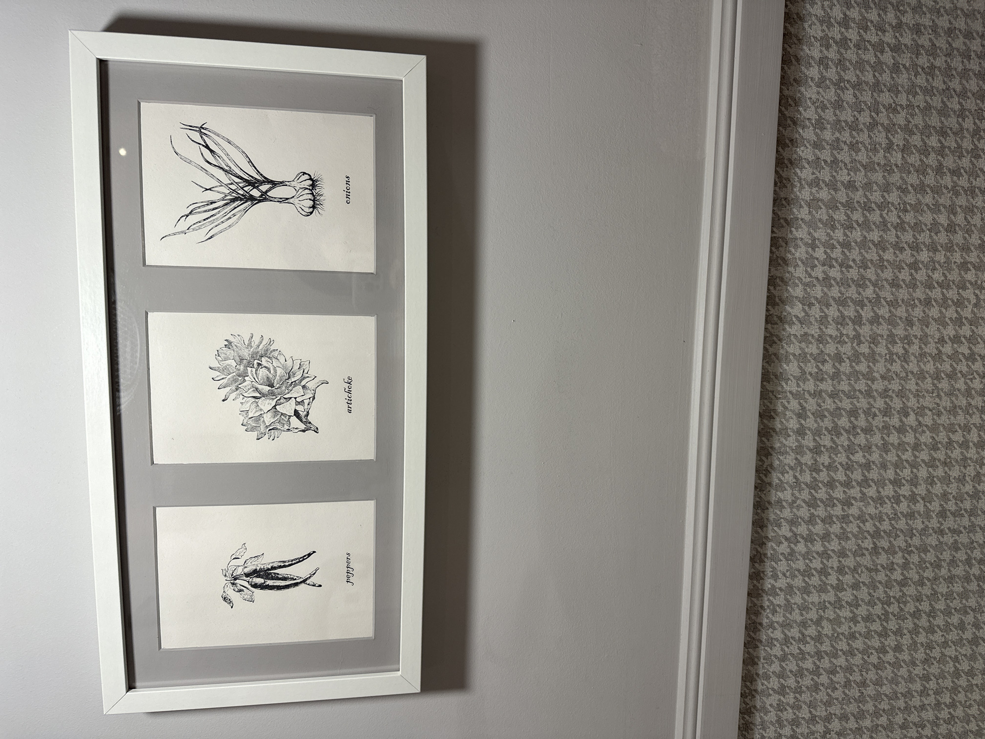 This is a close-up, eye-level, horizontal shot of a framed triptych art piece hanging on a light gray wall. The frame is white and appears to be made of wood or plastic. The triptych consists of three black and white botanical illustrations, each on a separate card. The illustrations are of vegetables, with text labels to their right.
The top illustration depicts a bunch of onions with their roots and green tops. The text label reads "onions".
The middle illustration shows an artichoke. The text label reads "artichoke".
The bottom illustration features a cluster of chili peppers. The text label reads "peppers".
The artwork is mounted on a gray mat board, which is enclosed within the white frame. The wall to the left of the artwork is a solid light gray, while the wall to the right features a gray and white houndstooth pattern. A white trim molding runs vertically along the right edge of the solid gray wall, separating it from the patterned wall.
The lighting is soft and even, suggesting an indoor setting during the daytime. There are no people or animals visible in the image. The overall mood is calm and decorative, likely intended for home or office decor. The context of Minsk, Belarus, is noted but not visually evident within the image itself.