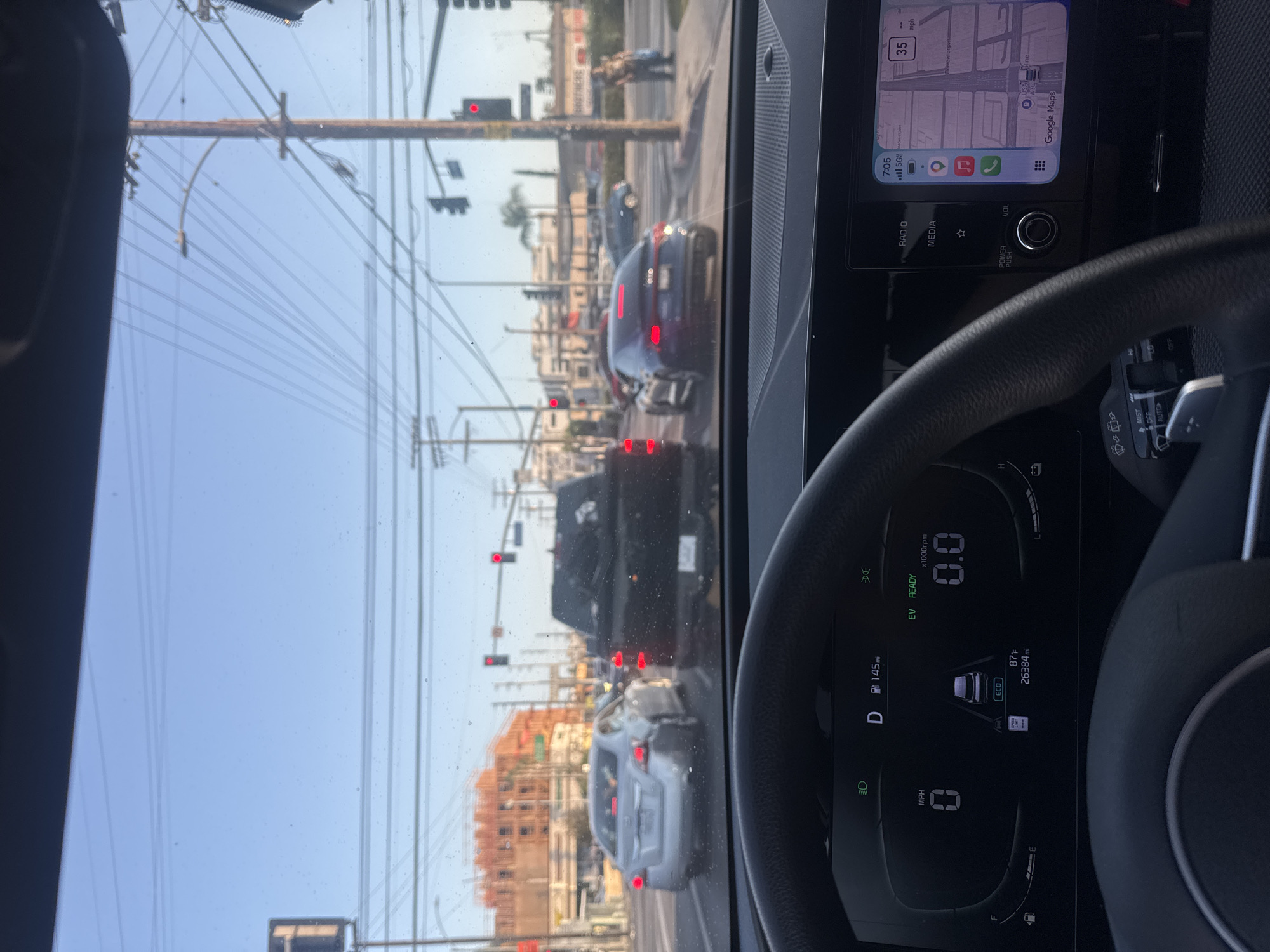 The image, taken from the perspective of a driver inside a car, shows a view through the windshield at an intersection in Los Angeles, California. The time is 7:05 AM. The sky is clear and blue. Several cars are stopped at the red traffic lights. A silver sedan is in the left lane, a black pickup truck is in the center lane, and a dark purple sports car is in the right lane. In the background, there is a multi-story building under construction. Traffic lights and utility poles with numerous wires are visible throughout the scene. The car's dashboard displays the current speed as 0 MPH, the gear as 'D', and the outdoor temperature as 87°F. The infotainment screen shows a navigation map and the current time. A person is visible on the sidewalk to the right of the intersection, holding a blue bag.