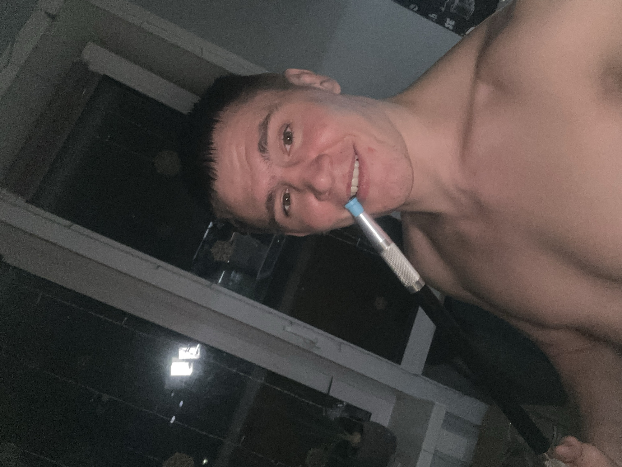 A shirtless man is holding a hookah mouthpiece in his mouth. He is looking at the camera and smiling. The setting is a dimly lit room, possibly in an apartment, with a window in the background. Without further information, determining the exact location in Perm, Russia is not possible.
