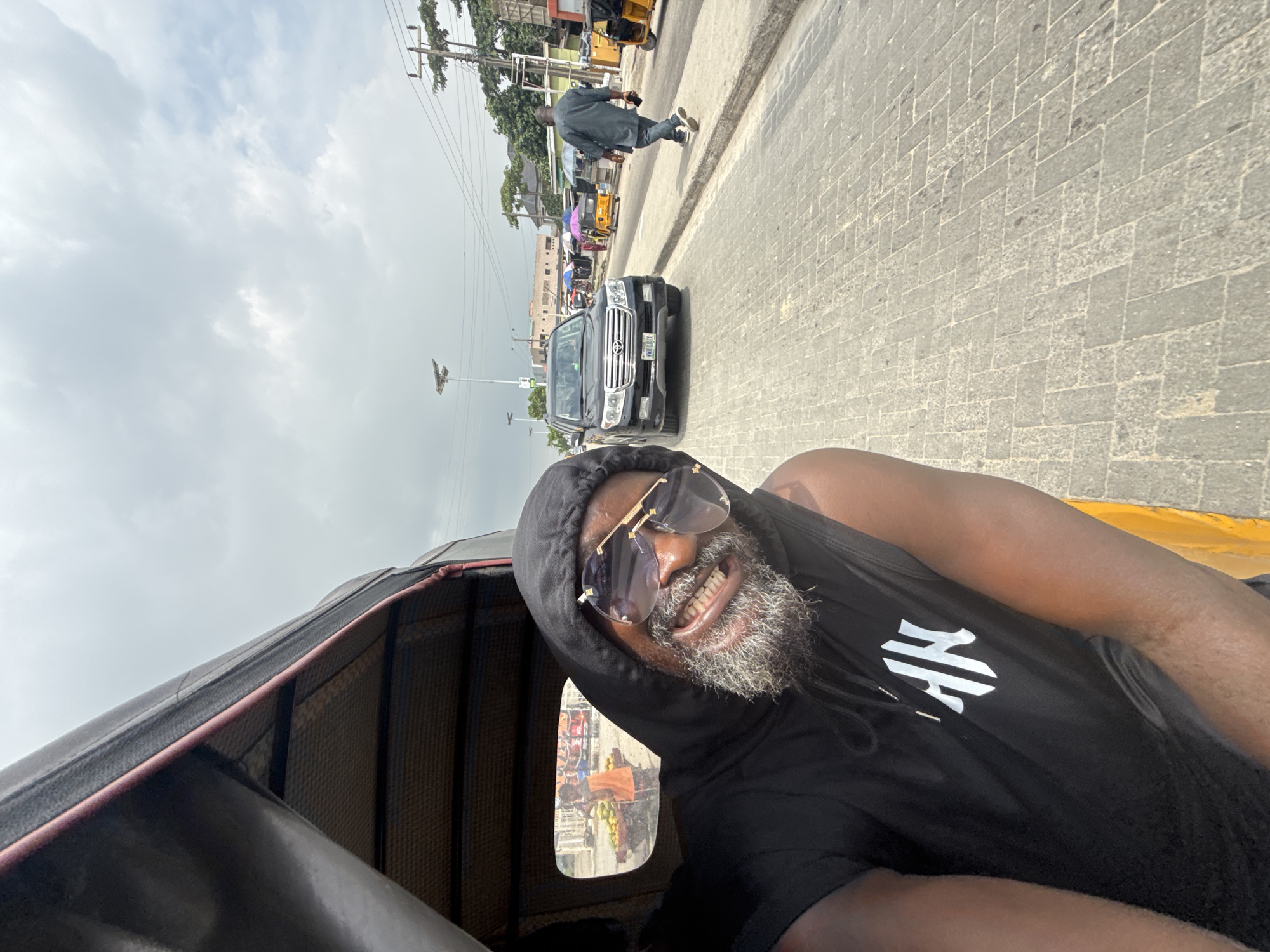 A man with a grey beard, wearing sunglasses and a black hoodie, is taking a selfie from inside a motorized rickshaw. The man is smiling broadly, showing his teeth. The rickshaw is on a paved street, and in the background, there are other vehicles, including a black SUV and several yellow motorcycles. Buildings and trees line the street, and there are power lines overhead. The sky is cloudy. The scene appears to be in Ado, Nigeria. A man in a dark shirt and blue jeans is walking on the sidewalk. There is some text on the man's hoodie, but it is not clearly legible.