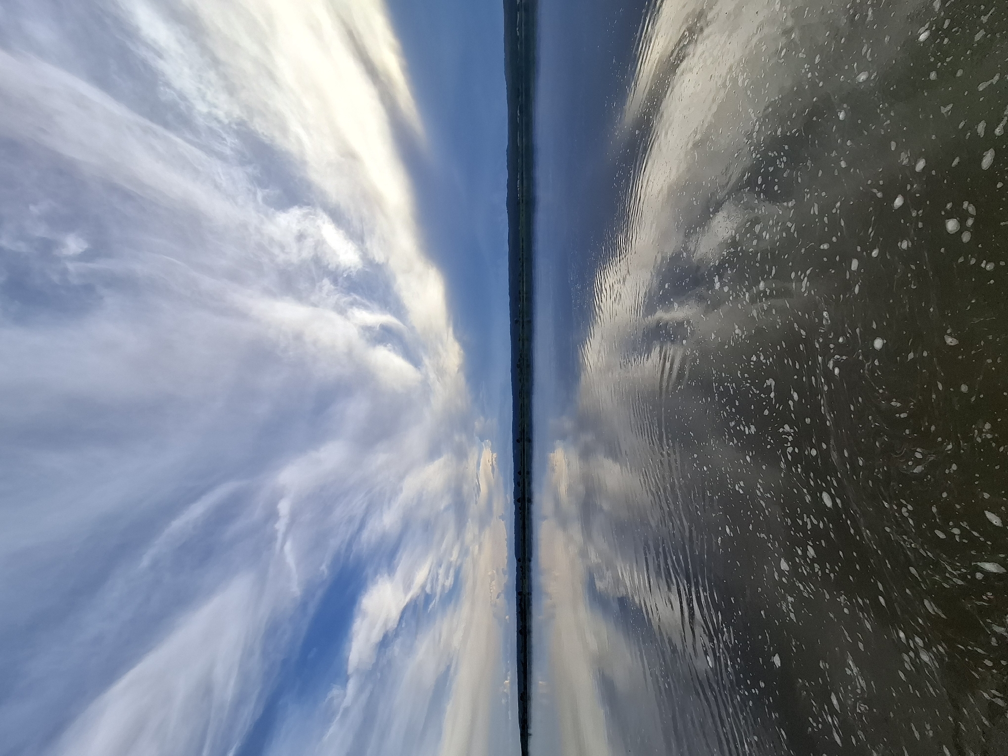 The image captures a wide-angle view of a serene body of water, likely a river or lake, in the city of Malaya Sazanka, Russia. The water's surface is calm, reflecting the dramatic sky above. Wispy, white clouds streak across a gradient of blue to darker, stormy hues in the sky. These clouds are mirrored on the water's surface, creating an impression of depth and vastness. The shoreline is a distant, dark silhouette, suggesting a natural landscape. In the foreground, the water is darker and shows signs of slight ripples, with scattered white foam or bubbles dotting the surface. There are no people or man-made structures prominently visible. The overall atmosphere is one of natural tranquility and expansive sky.