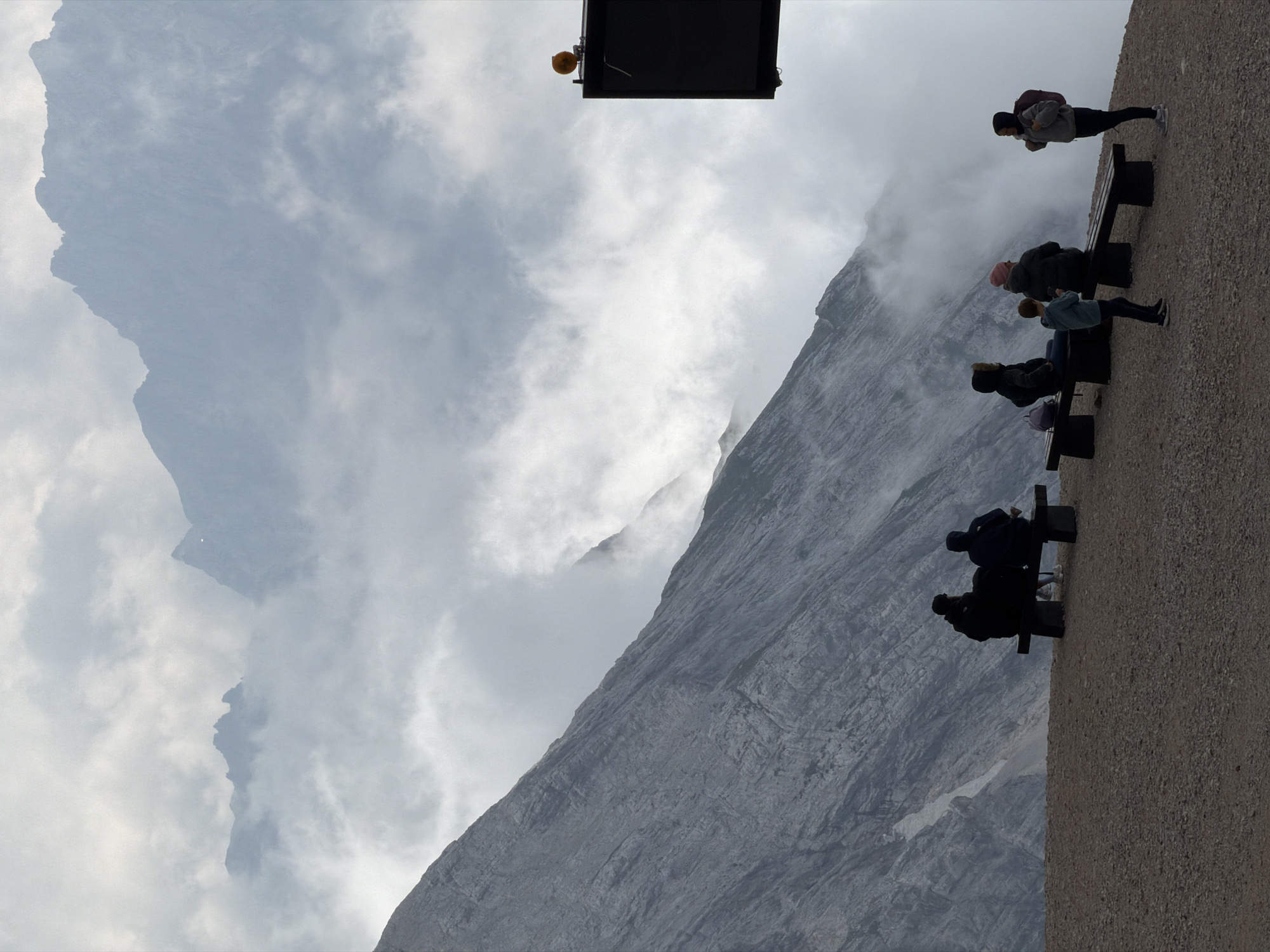 The image depicts a scenic view from a high altitude, likely the Zugspitze, Germany's highest peak, near Garmisch-Partenkirchen. In the foreground, several people are seated and standing on benches. To the left, two individuals are seated on a bench, facing away from the camera. Further to the right, three more individuals, two adults and a child, are seated on another bench. To the far right, a person stands facing the others, with a backpack on. A dark sign with an orange light is visible on a pole in the upper right corner.
The background is dominated by a steep, rocky mountain slope on the left, shrouded in mist and clouds. The upper part of the image reveals majestic mountain ranges receding into the distance, partly obscured by atmospheric haze and clouds, suggesting a cool, potentially damp, mountain environment. The ground in the foreground is covered with gravel and small stones. The overall atmosphere is serene and contemplative, with the people appearing to enjoy the dramatic mountain panorama.
