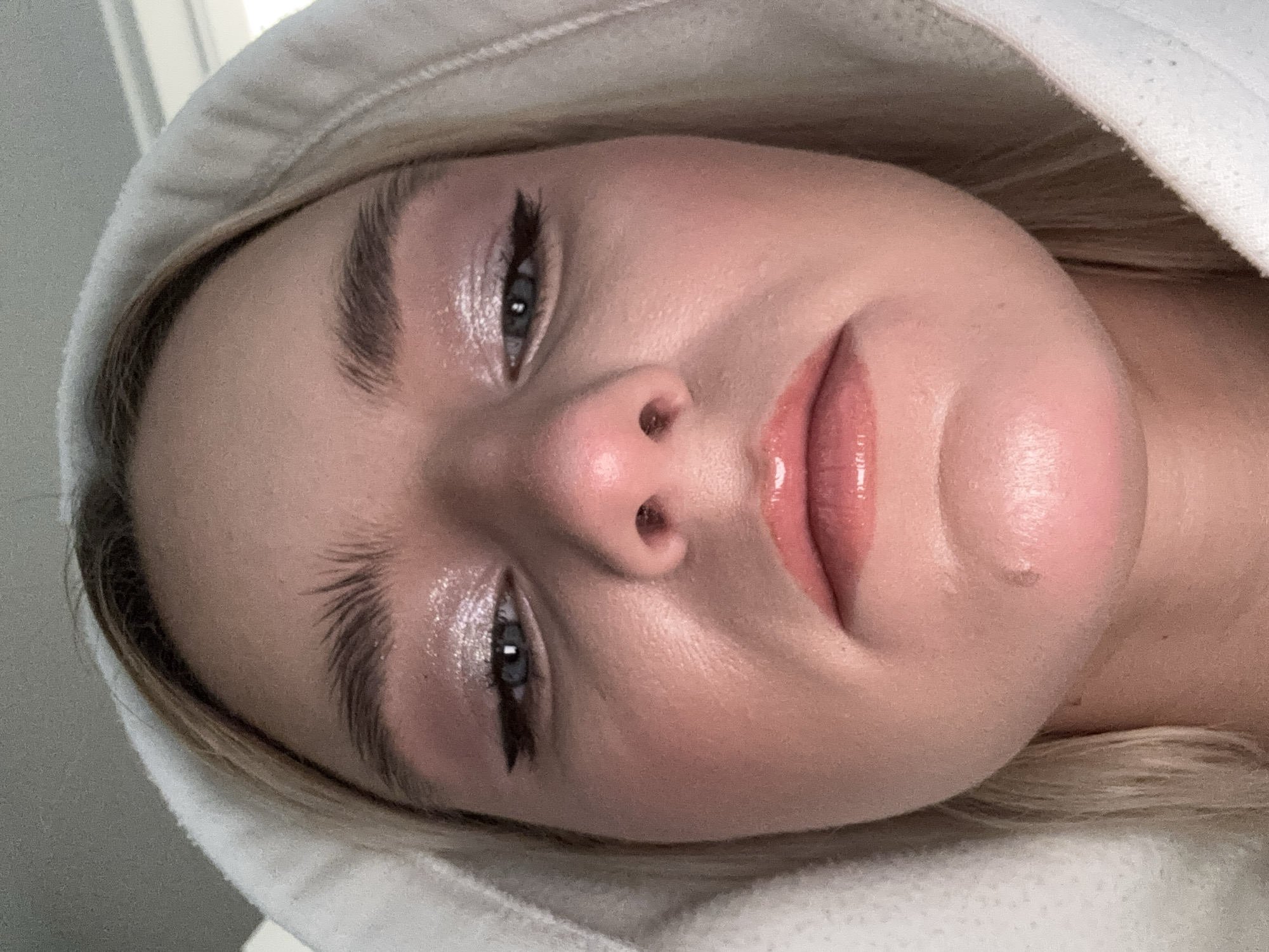 A close-up, rotated image depicts a fair-skinned person wearing a white hooded sweatshirt. The individual has prominent eyebrows, light-colored eyes, and glossy lips. Eyeliner is visible on the upper eyelids. The background is indistinct and primarily light-toned. There is a blemish visible on the right cheek, near the corner of the mouth. The location is not identifiable within the image. The context of "Unknown, Russia" is irrelevant.