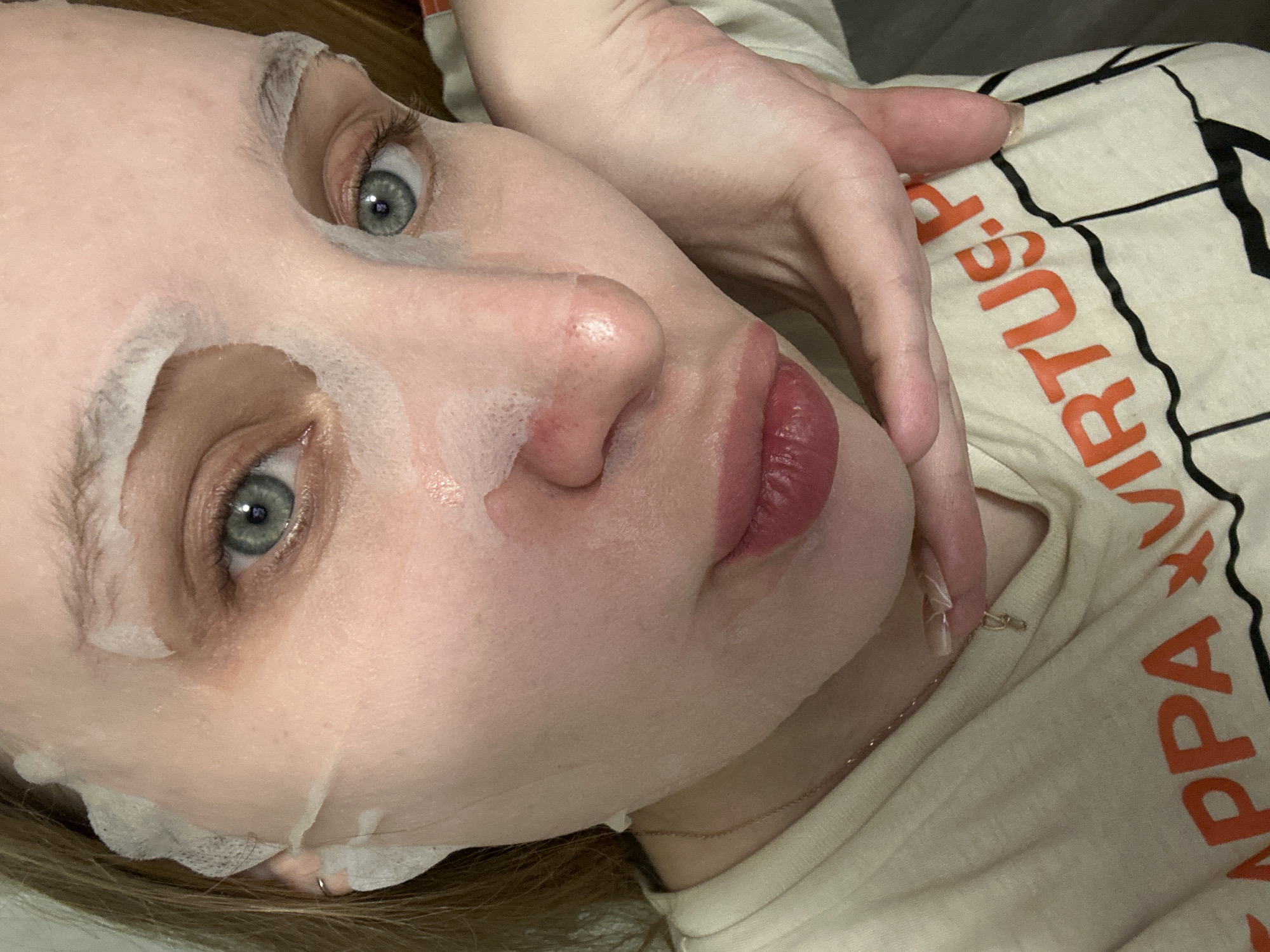 A woman is reclining and facing upward, with a sheet-type face mask applied to the left side of her face, while her hand is near the right side of her face. She has fair skin and light-colored eyes. She is wearing a light-colored shirt with orange lettering that includes the word "VIRTUES." A thin gold necklace is also visible around her neck. The photo's location is not discernible and cannot be identified as Chaikovsky, Russia, or any other specific location.