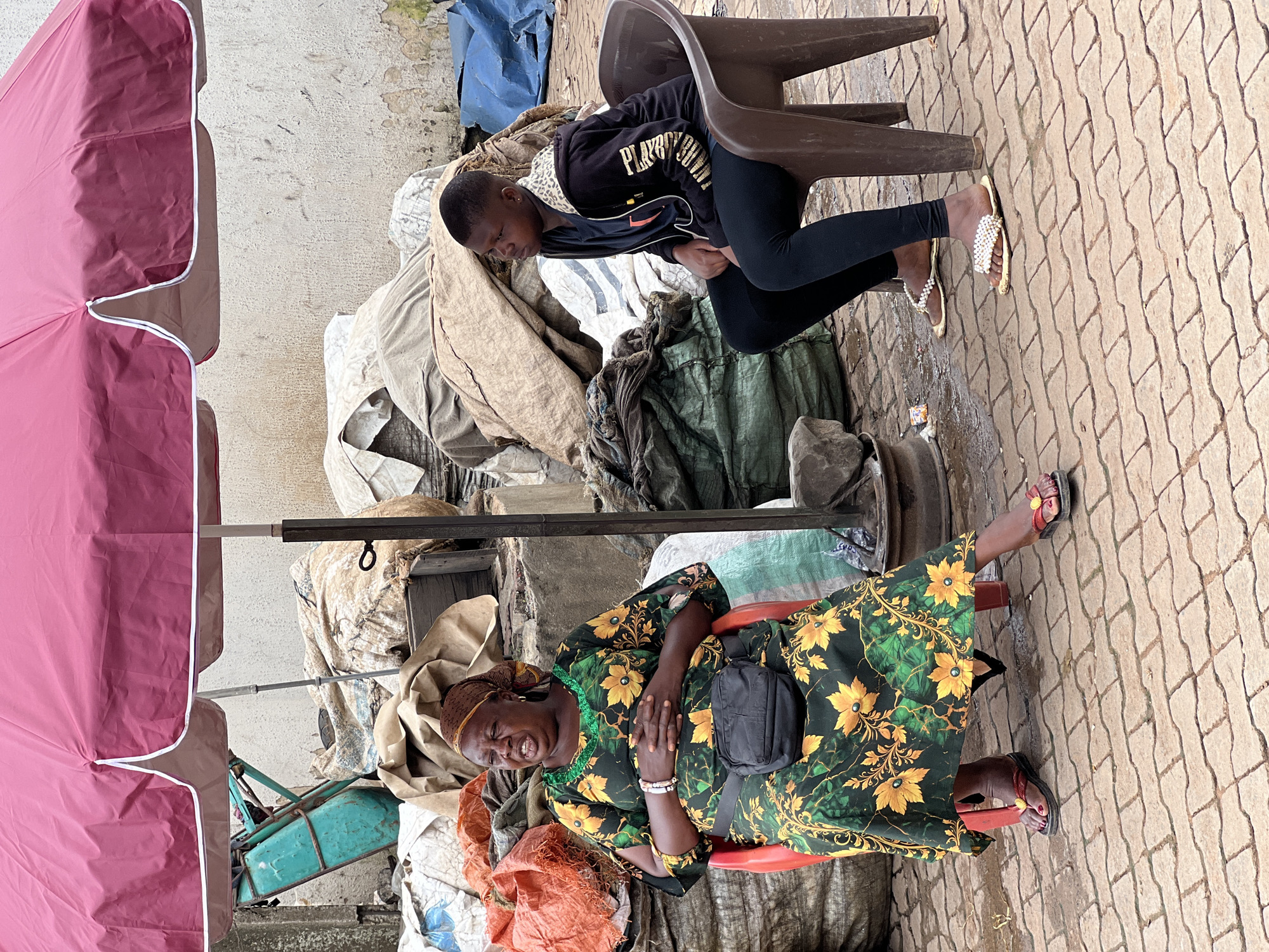 Two individuals are seated under a large, pink umbrella at an outdoor market in Nkpor, Nigeria. The person on the left is a woman wearing a green and yellow floral dress, a headscarf, and a black fanny pack. She is sitting on a red chair with her hands clasped in her lap and her mouth open as if speaking. The person on the right is a younger individual, possibly a girl, wearing a black hoodie, black leggings, and sandals adorned with pearls. She is seated on a brown plastic chair and looking to her right. The ground is paved with interlocking bricks, and the area is filled with large sacks and bundles of materials, suggesting a marketplace or storage area. A teal-colored cart is visible behind the woman.