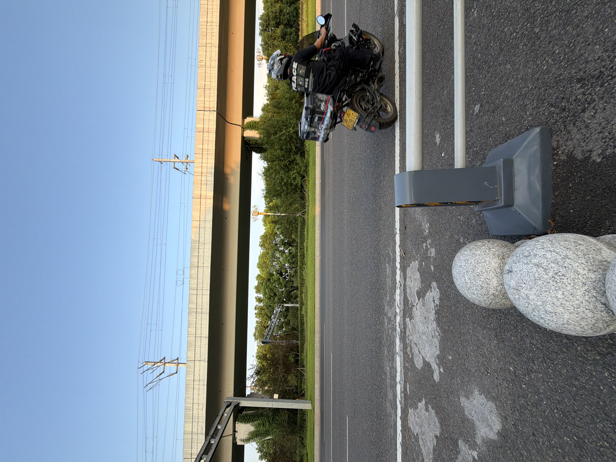 This image captures a street scene in Jiangjin, China, under a clear blue sky at what appears to be either sunrise or sunset, given the warm lighting on the elevated structure. In the foreground, two large, round, speckled granite spheres sit on the asphalt road, next to a gray barrier with horizontal white bars. A motorcycle is being ridden on the road to the right of the frame. The rider, wearing a helmet and a black shirt with the word "PEACE" printed on it, is facing away from the camera. The motorcycle is black and has a large gray storage box on the back, with various logos and designs, including "GS". The license plate is yellow and black with Chinese characters and the number "B2B10". An elevated concrete overpass, supported by pillars covered in green foliage, stretches across the upper portion of the image. Power lines are visible strung along poles above the overpass. Behind the road and the overpass, a dense line of green trees forms the background, with some taller structures or poles interspersed within them. The overall environment suggests an urban or suburban setting with significant infrastructure.