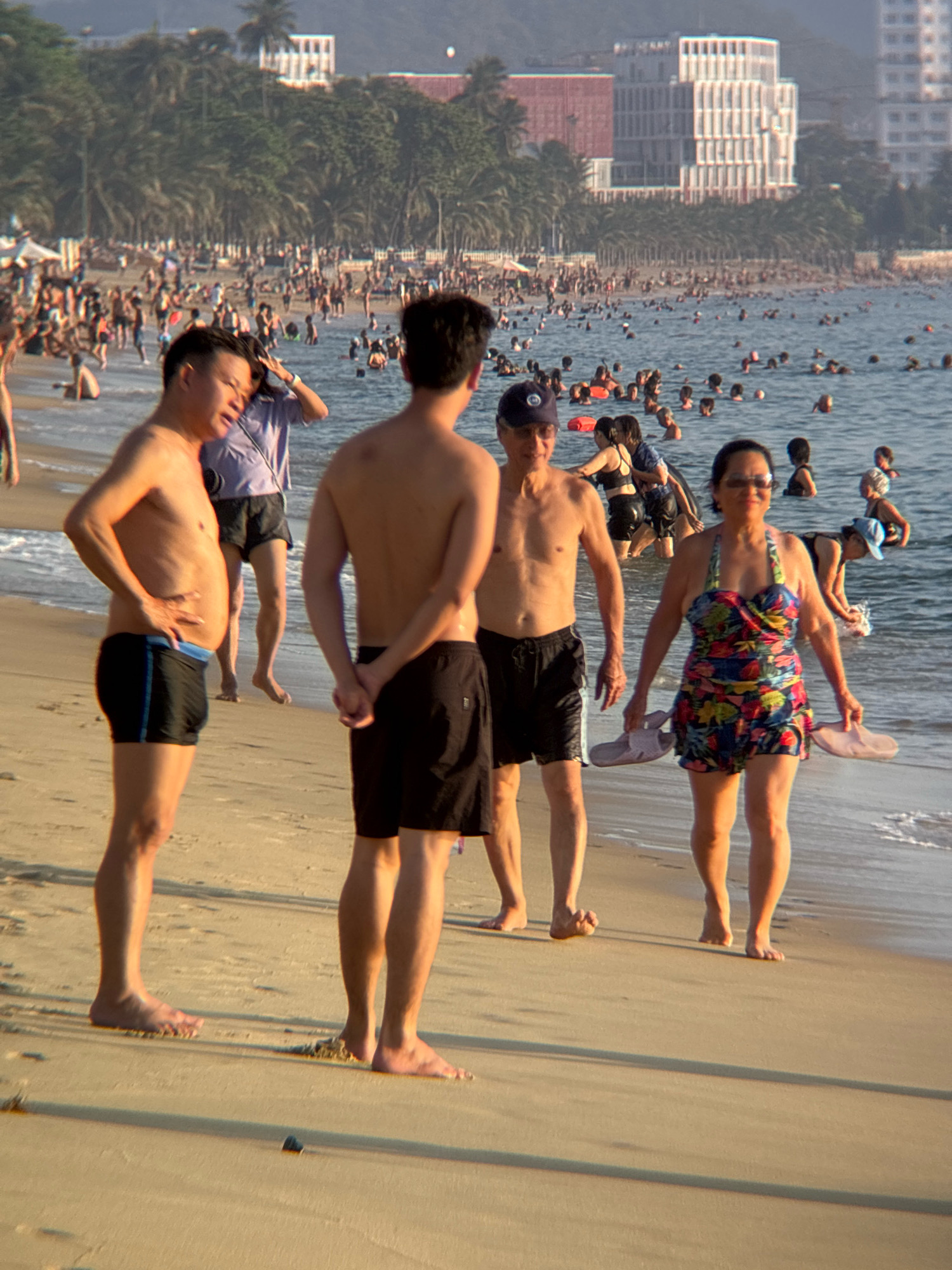 The image depicts a crowded beach scene in Nha Trang, Vietnam. In the foreground, three men and one woman are positioned on the sandy shore. The man on the far left is facing away from the camera, wearing black swim trunks with a blue stripe. Next to him, a man is facing the camera, showing his torso and hands on his hips, also wearing black swim trunks. To his right, another man is facing away, wearing black swim trunks. The woman in the foreground is facing the camera, wearing a colorful floral swimsuit and holding two pink flip-flops. She is walking towards the viewer. In the background, the ocean is filled with numerous people swimming and wading. Beyond the water, a shoreline is visible with palm trees and buildings, characteristic of Nha Trang's coastal landscape. The lighting suggests it is late afternoon or early evening, with long shadows cast across the sand. The atmosphere is lively and summery, with many individuals enjoying the beach and ocean.
