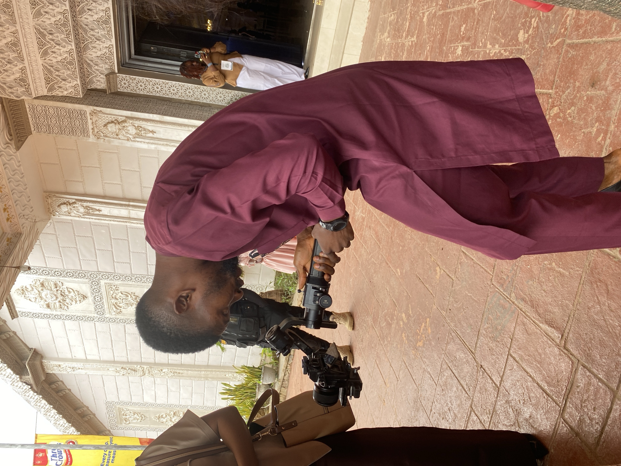 The image shows a man in a maroon traditional outfit operating a camera gimbal in front of an ornate building. The building has white walls with intricate decorative patterns and a large entrance. The ground is paved with reddish-brown tiles. In the background, other individuals are visible, including a woman in a brown top and white pants near the entrance and another person in dark tactical gear. A yellow banner with some text is visible on the left side of the image. The overall scene suggests an event or gathering is taking place in a decorated outdoor setting in Jos, Nigeria.