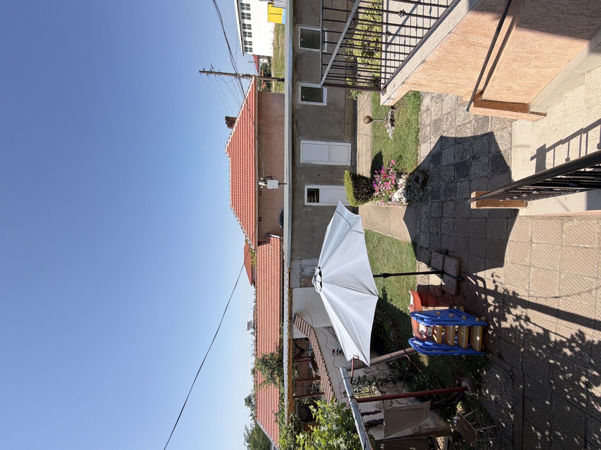 The image depicts a sunny, outdoor scene in the city of Dolni Dabnik, Bulgaria. The viewpoint is from an elevated position, looking down into a backyard area. A light-colored umbrella is open in the center of the frame, casting a shadow. To the left of the umbrella, a blue and yellow plastic children's slide is visible. A small bed of pink flowers is situated on the grass to the right of the slide. Beyond the backyard, a single-story building with a red-tiled roof is prominent, with concrete walls and white doors and windows. Further in the background, a larger white building with a multi-paned facade can be seen, along with utility poles and overhead wires. The ground is paved with square tiles in some areas and covered with grass in others. A black metal railing runs along the edge of the elevated vantage point. The sky is clear and bright blue, indicating fair weather.