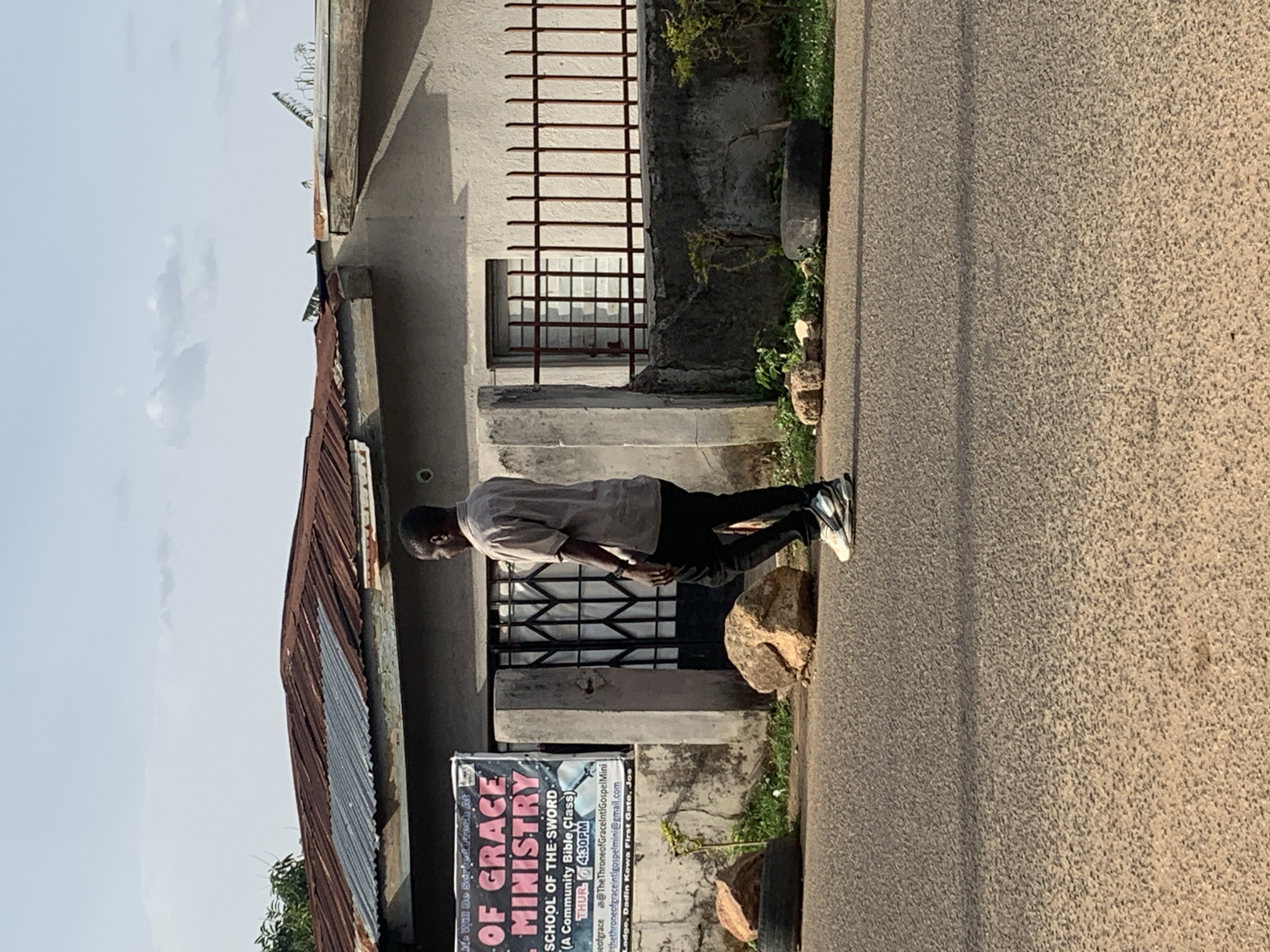 A man stands on a rock along a street in Jos, Nigeria, in front of a building bearing a sign for the "Of Grace Ministry, School Of The Sword, A Community Bible Class." The class is held Thursdays at 4:30 PM, and the sign lists social media accounts and an address: Dadin Kowa First Gate, Jos. The building is made of cement or stucco, with barred windows and a corrugated metal roof. The man is wearing a light-colored shirt, dark pants, and light-colored shoes with dark accents.