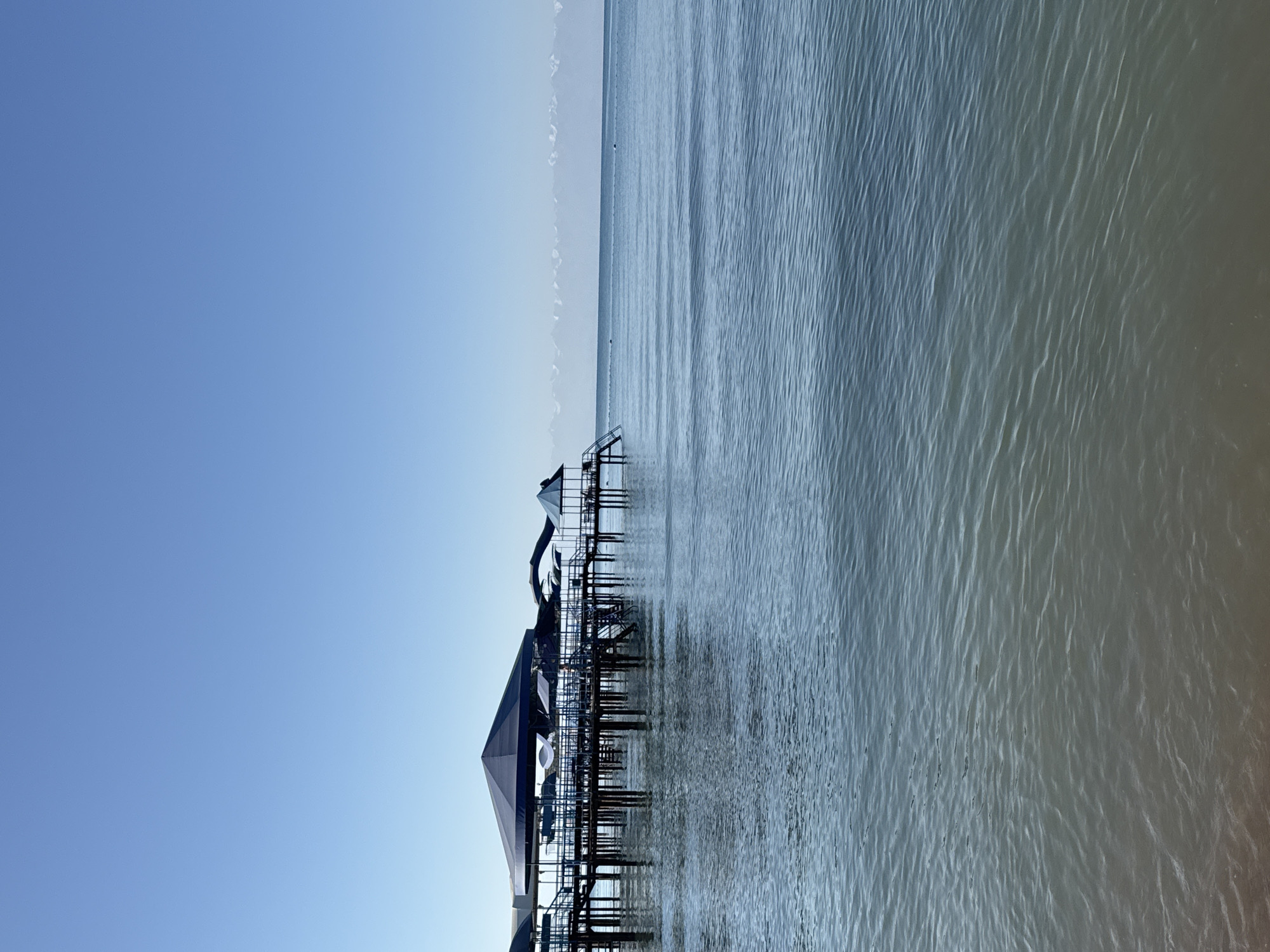 A pier extends into a calm, blue body of water under a clear, bright blue sky. The pier is supported by numerous vertical poles and has several structures on it, including large, dark blue canopies, smaller shelters, and what appear to be slides or diving platforms. A distant, snow-capped mountain range is visible on the horizon, separating the water from the sky. The water exhibits gentle ripples, and the bottom is visible in the shallower areas near the foreground, revealing a sandy or pebbly shore. No people are visible in the scene. The location is Sary-Oy, Kyrgyzstan.