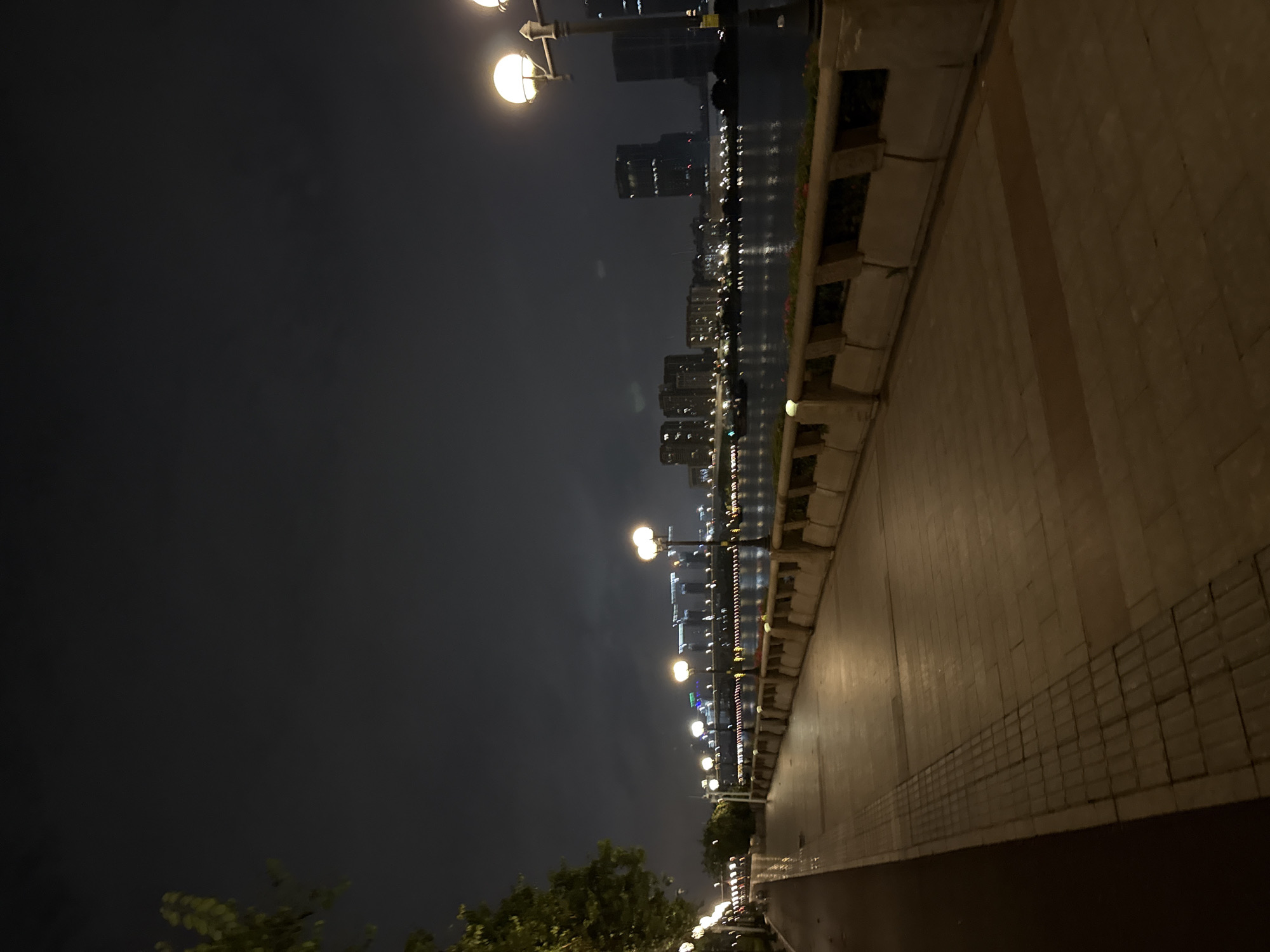 A nighttime view of a waterfront promenade in Tianhe District, China, reveals a paved walkway bordered by a low wall with planters. Streetlights with glowing globes illuminate the path, casting warm light on the textured paving stones and a darker, red-hued path to the left. Across the water, a city skyline is visible, featuring illuminated buildings and bridges. A few trees are present on the left side of the promenade, their leaves dark against the night sky. The water itself reflects the distant city lights. The scene is tranquil and deserted.