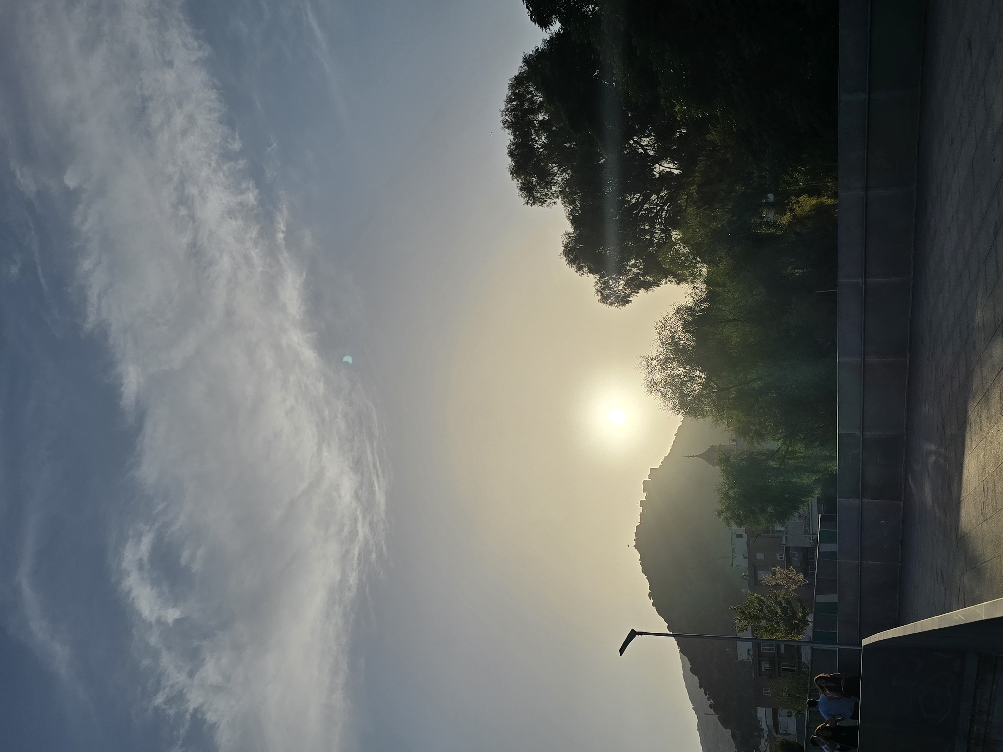 The image shows a view of the city of Jaén, Spain, looking towards the Castillo de Santa Catalina. The sun is low in the sky, casting a warm glow and creating a hazy atmosphere. The Castillo, perched atop a hill, is silhouetted against the bright sky, with a cross visible on its highest point. Some structures, possibly part of the castle or surrounding buildings, are also discernible.
In the foreground, there is a paved area and a low wall. A street lamp with a modern design stands on the left side of the image, near the hill. Behind the wall, there are some trees, including a prominent cluster of dark green trees on the right, and lighter, more wispy trees in the middle ground. Buildings are visible on the hillside, interspersed with the trees.
Several people are standing near the wall in the middle left of the frame, appearing to be casually observing the scene. Their backs are mostly to the camera, and they are dressed in casual clothing. The sky is a light blue with scattered clouds, some of which are wispy and streaky, while others are more dense and grey. There is a noticeable lens flare or atmospheric distortion in the upper central part of the image. The overall impression is that of a late afternoon or early evening view of the city under a bright, setting sun.