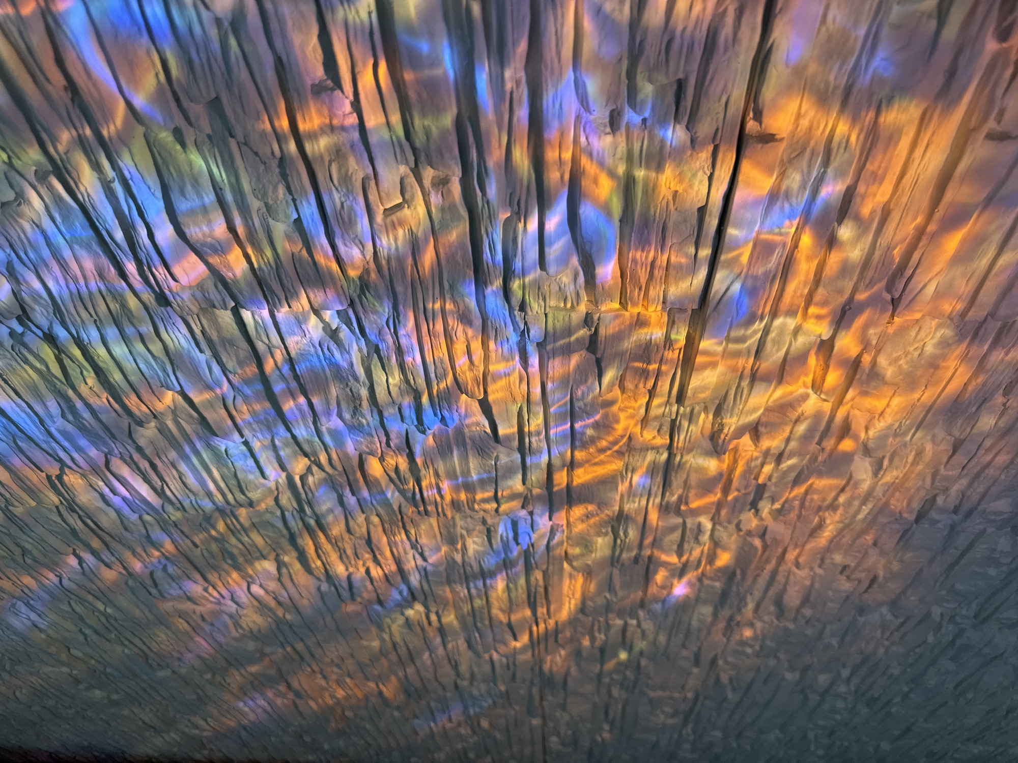The image presents a close-up view of a textured surface illuminated with a dynamic interplay of colors, predominantly orange, blue, and yellow. The texture appears to be composed of raised, vertical ridges or folds, casting shadows that accentuate the three-dimensional quality. The light filtering through these structures creates vibrant patterns, suggesting a natural light source refracted or reflected. The location is not specified; therefore, it is impossible to discern details about the city of Unknown, Unknown.