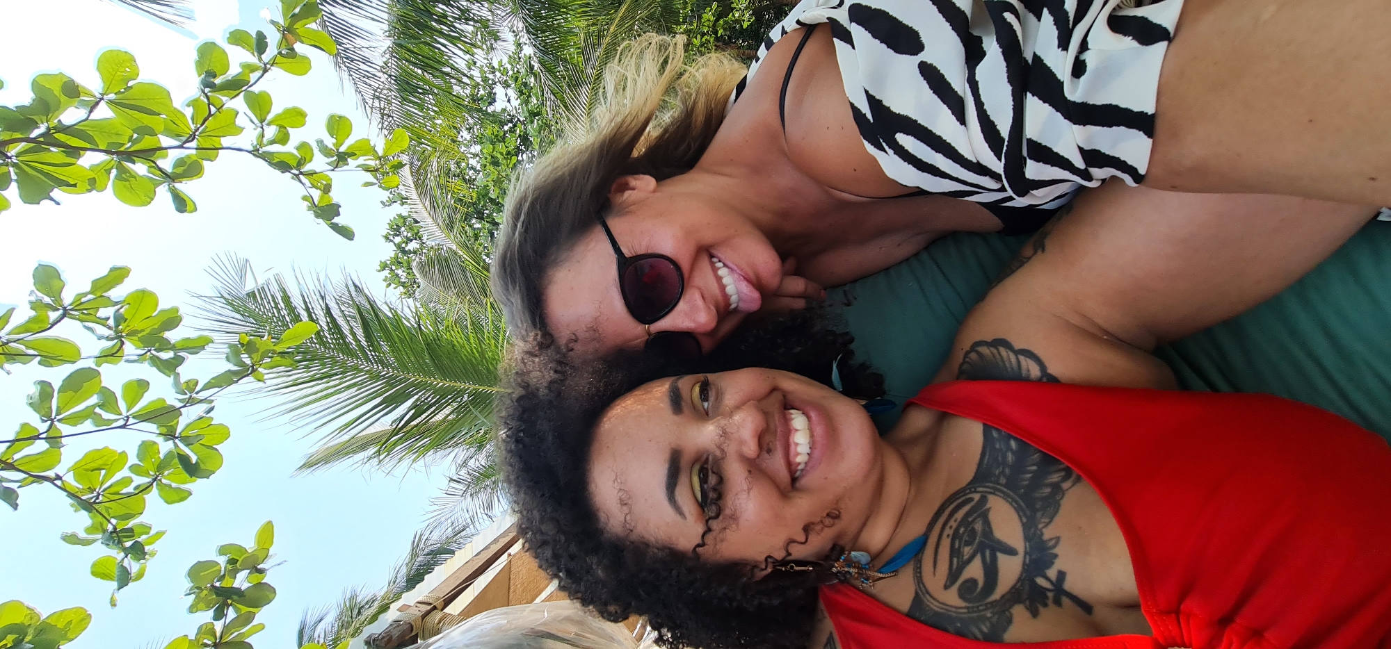 Two women pose closely for a photo under trees. One woman wears sunglasses and a black and white patterned top. The other woman has curly hair, a red top, and tattoos on her chest. They both smile. Palm and other foliage are visible behind them. The setting appears to be outdoors in a tropical location. The location in the city of Đà Nẵng, Vietnam, cannot be confirmed based solely on the image.