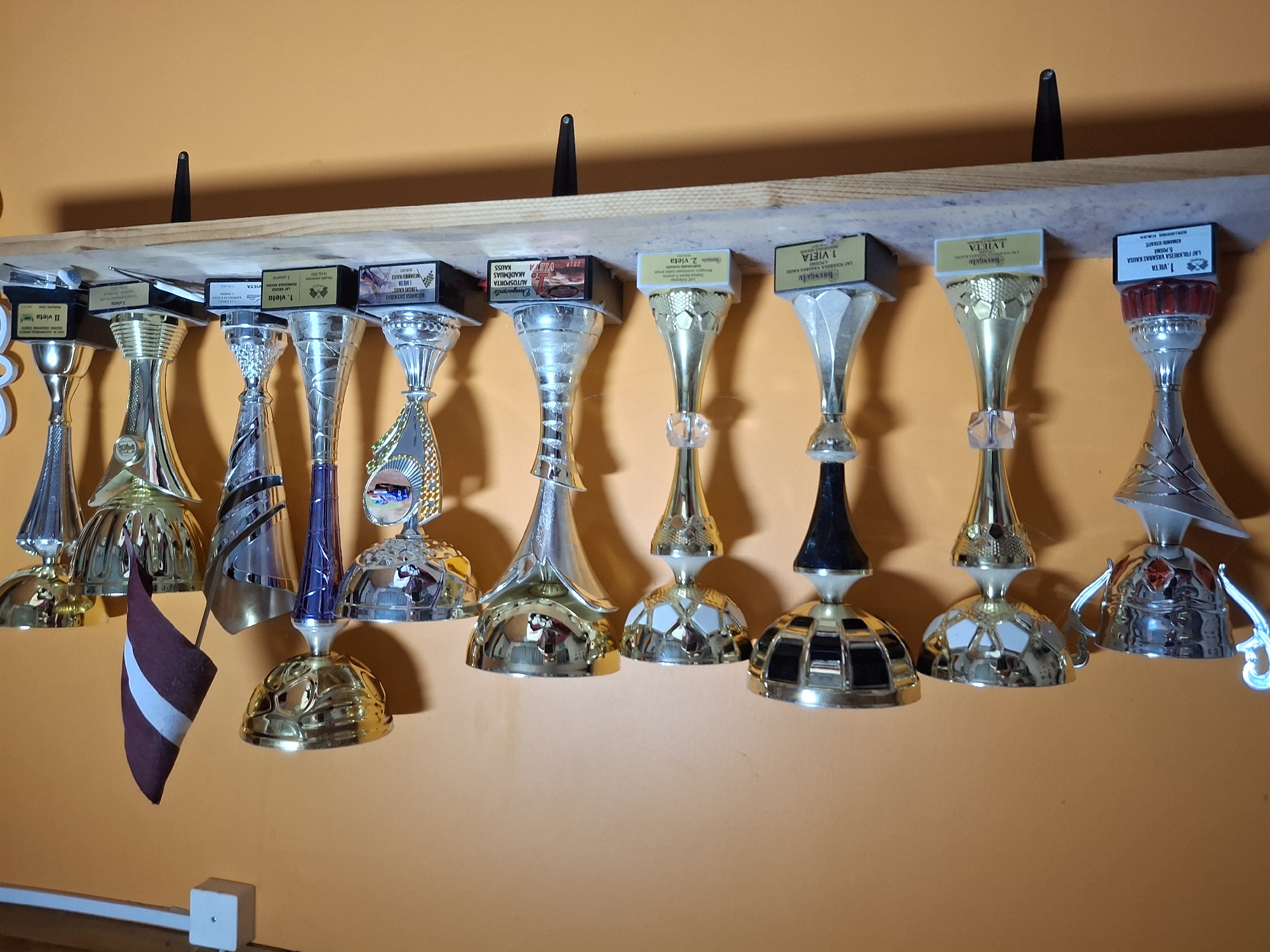 The image shows a shelf mounted on an orange wall, displaying a collection of trophies. There are at least eleven trophies of varying sizes and designs, mostly made of gold-colored metal and silver-colored metal, with some featuring black bases. A small Latvian flag is placed among the trophies on the right side of the shelf. The shelf itself is made of wood and is supported by dark brackets. On the wall above the shelf, there is a white electrical box. The overall setting appears to be indoors, possibly a home or an office, in the city of Ogre, Latvia. The lighting suggests an indoor environment, and the orange wall and wooden shelf are prominent environmental features. There are no people visible in the image.