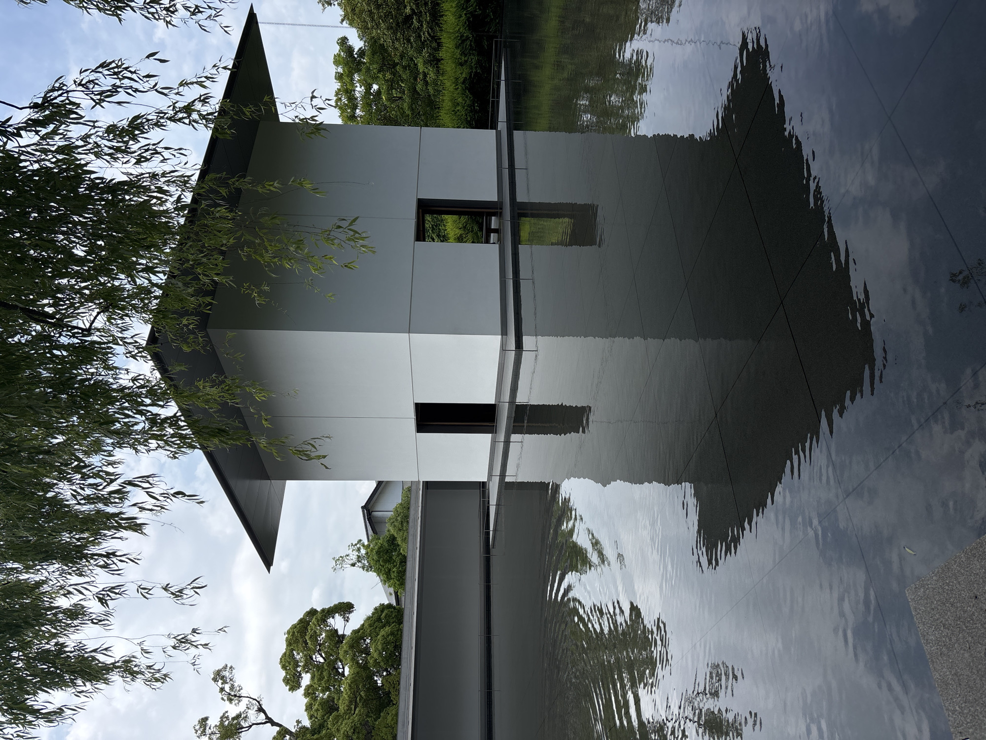 A minimalist, modern, white, cube-like building with a wide, dark grey overhanging roof stands centrally within a dark, still reflective pool of water under an overcast sky. The building features two dark, rectangular open doorways on the side facing the viewer, which are clearly reflected in the water below. The water's surface exhibits subtle ripples, particularly on the left side, slightly distorting the reflections of the sky and surrounding foliage. The building itself is situated on a dark, tiled platform that extends into the water.
In the upper portion of the frame, weeping willow branches with slender green leaves hang down, partially obscuring the building's roof and the sky. To the left of the building, a long, low, grey wall runs horizontally, behind which lush green trees with dense canopies are visible. Further in the background, a traditional Japanese-style dark tiled roof is discernable amongst the trees. To the right of the building, dense green bushes and trees form a continuous backdrop. The reflections in the water accurately mirror the building's white walls, dark doorways, and the overhead sky and willow branches. No people are visible in the scene. This architectural scene is characteristic of the D.T. Suzuki Museum in Kanazawa, Japan.