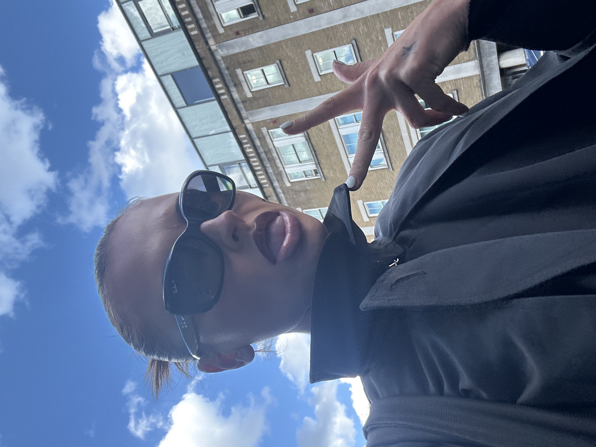 A woman with blonde hair pulled back, wearing dark sunglasses, and dark clothing, poses for a selfie outdoors. Her tongue is slightly out, and her right hand is held up, forming a peace sign. She has light-colored nail polish on her fingers. Behind her, a beige brick building with multiple windows and a light-colored modern addition on top is visible against a bright blue sky with scattered white clouds. The setting appears to be urban.