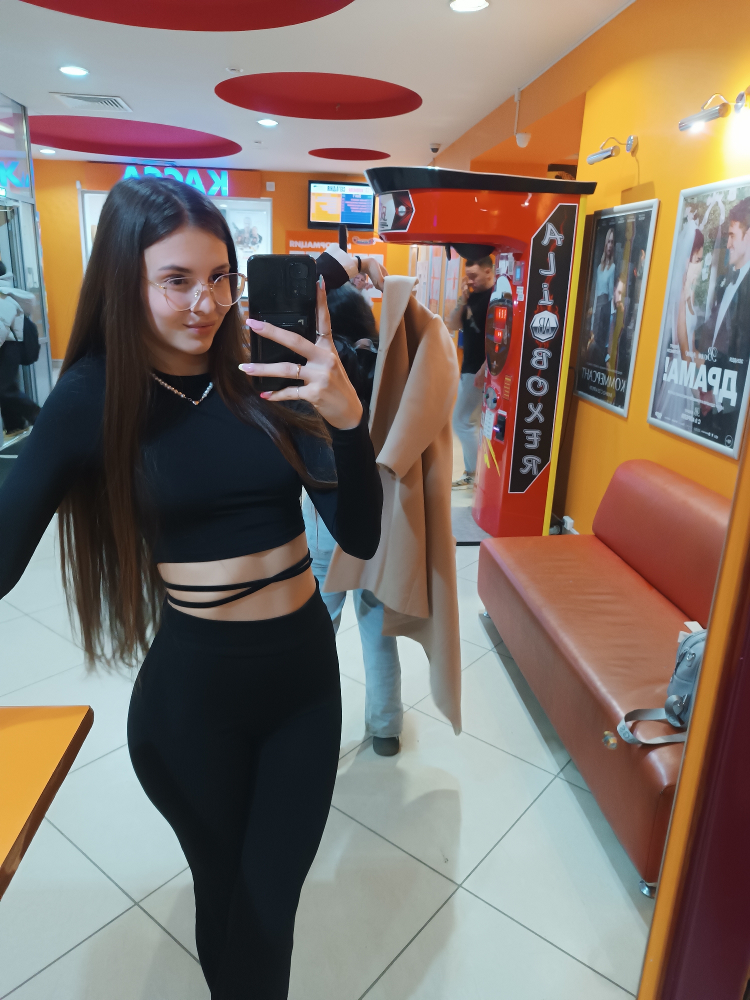 A young woman is taking a selfie in a mirror. She is wearing a black long-sleeved crop top and black leggings. She is also wearing glasses and a necklace. The background of the image appears to be a public space, possibly a game room or arcade. There's a punching bag machine, a bench seat, and framed posters on the wall. Two people are in the background near the punching bag machine, one holding a light-colored coat. The floor is tiled.