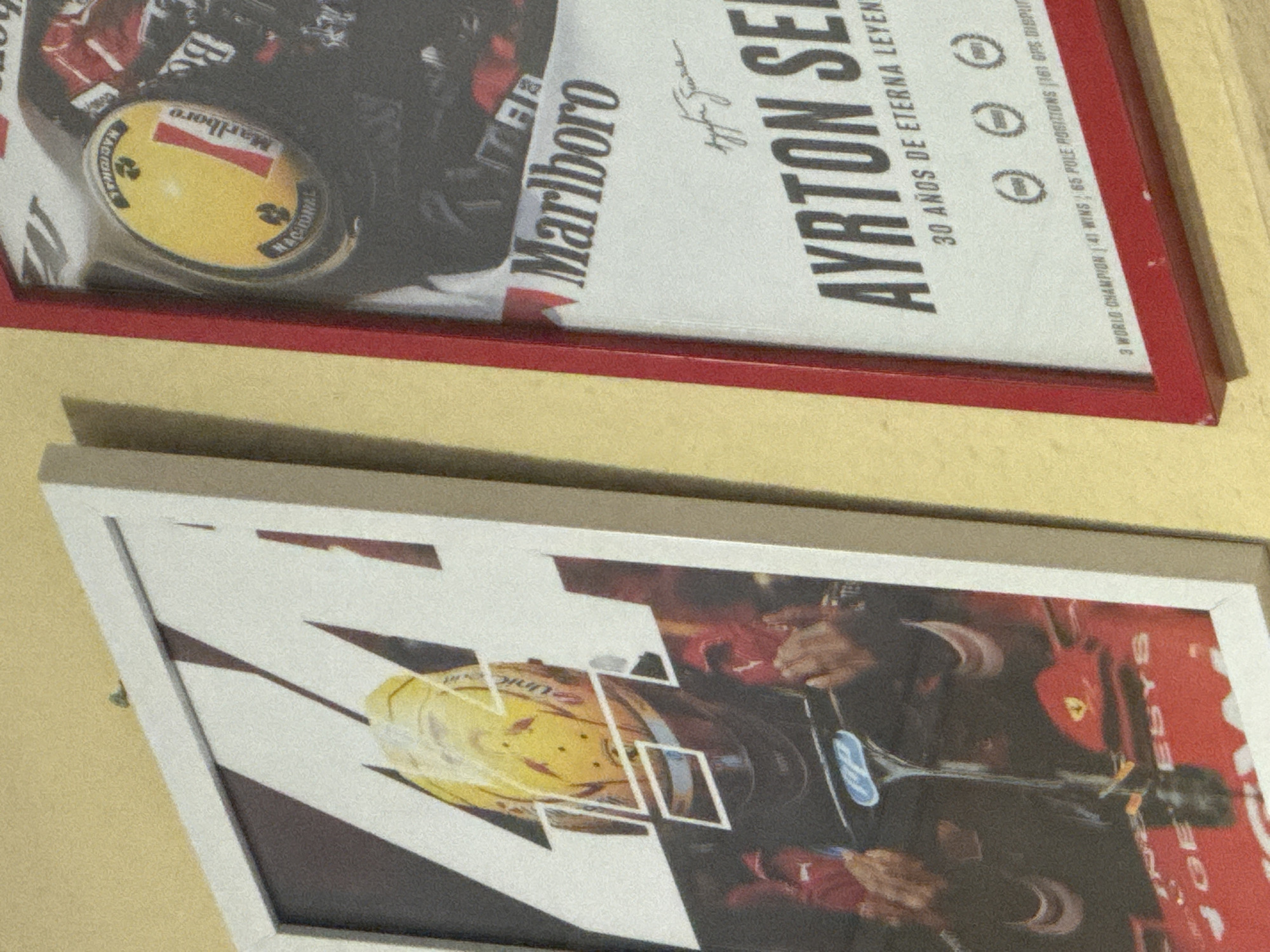 The image shows two framed posters hanging on a yellow wall. The poster on the left is framed in white and features a close-up of a racing helmet with the number "44" superimposed. The helmet is yellow with red and white accents. Below the helmet, a portion of a red race car is visible, with the text "JOE ESYS" printed on it. The poster on the right is framed in red and displays a portrait of Ayrton Senna. The top of the poster shows a yellow helmet with "Marlboro" branding. Below the portrait, the text reads "AYRTON SENNA 30 AÑOS DE ETERNA LEYENDA" and statistics such as "3 WORLD CHAMPION | 41 WINS | 65 POLE POSITIONS | 161 GPS DISPUTED." The lighting suggests an indoor environment, possibly a home or a collector's display.