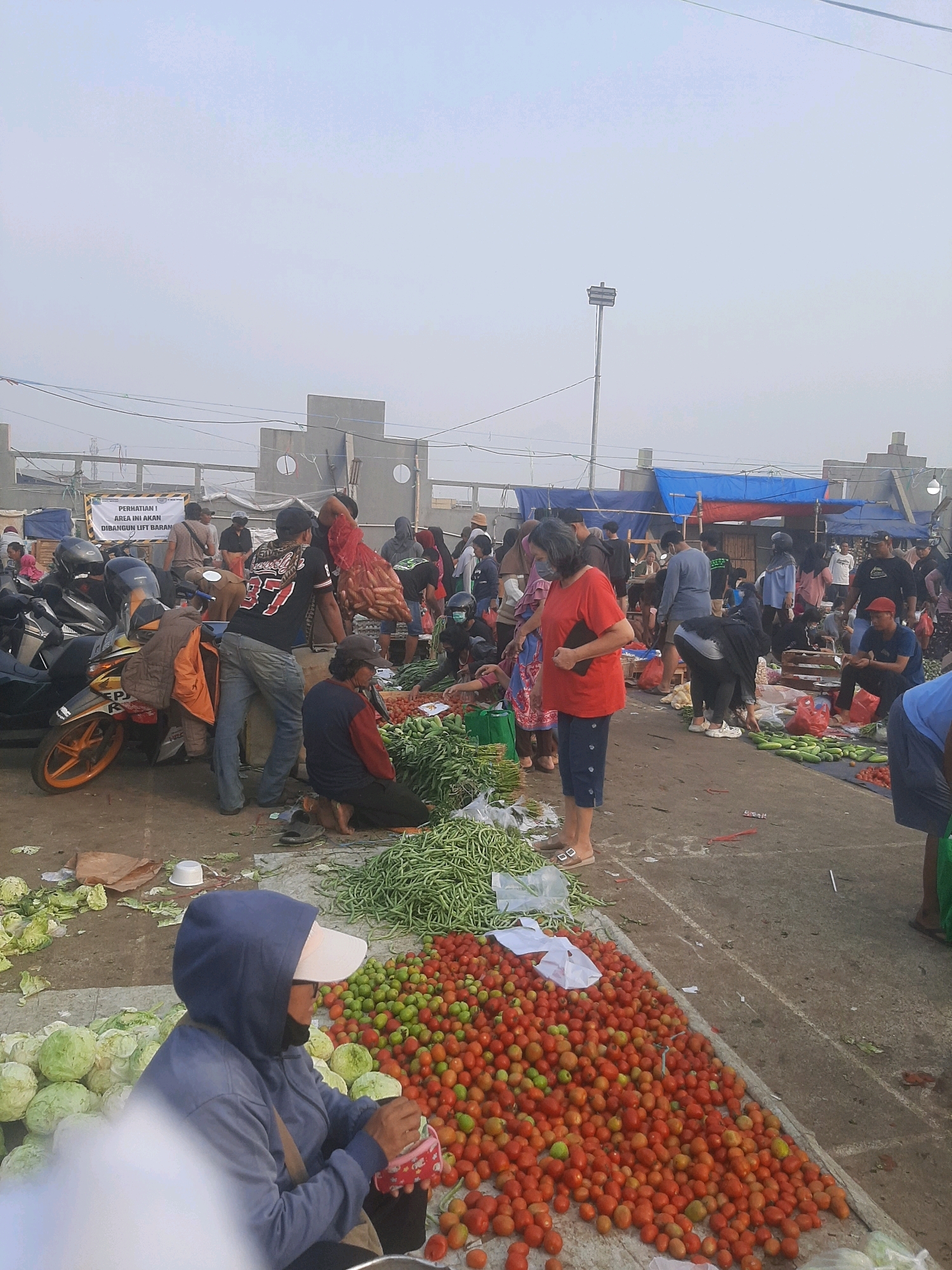 This is a wide-angle, eye-level shot of an outdoor market in Bekasi, Indonesia. The sky is overcast, creating a diffused light. In the foreground, large piles of fresh produce, including green beans, tomatoes, and cabbages, are displayed on the ground, some in woven baskets or plastic bags. Several people are present, engaged in various activities. One person in a blue hooded sweatshirt and a light-colored baseball cap is sorting through produce. Another person in a red t-shirt and blue pants is holding a phone. In the middle ground, more people are browsing and selling goods. Motorcycles are parked on the left side of the image, and some appear to be in use. In the background, there are temporary stalls covered with tarps of blue and red. A sign is visible with Indonesian text, and some concrete structures suggest ongoing construction or development in the area. The ground surface is concrete and appears somewhat dirty, with some scattered debris.