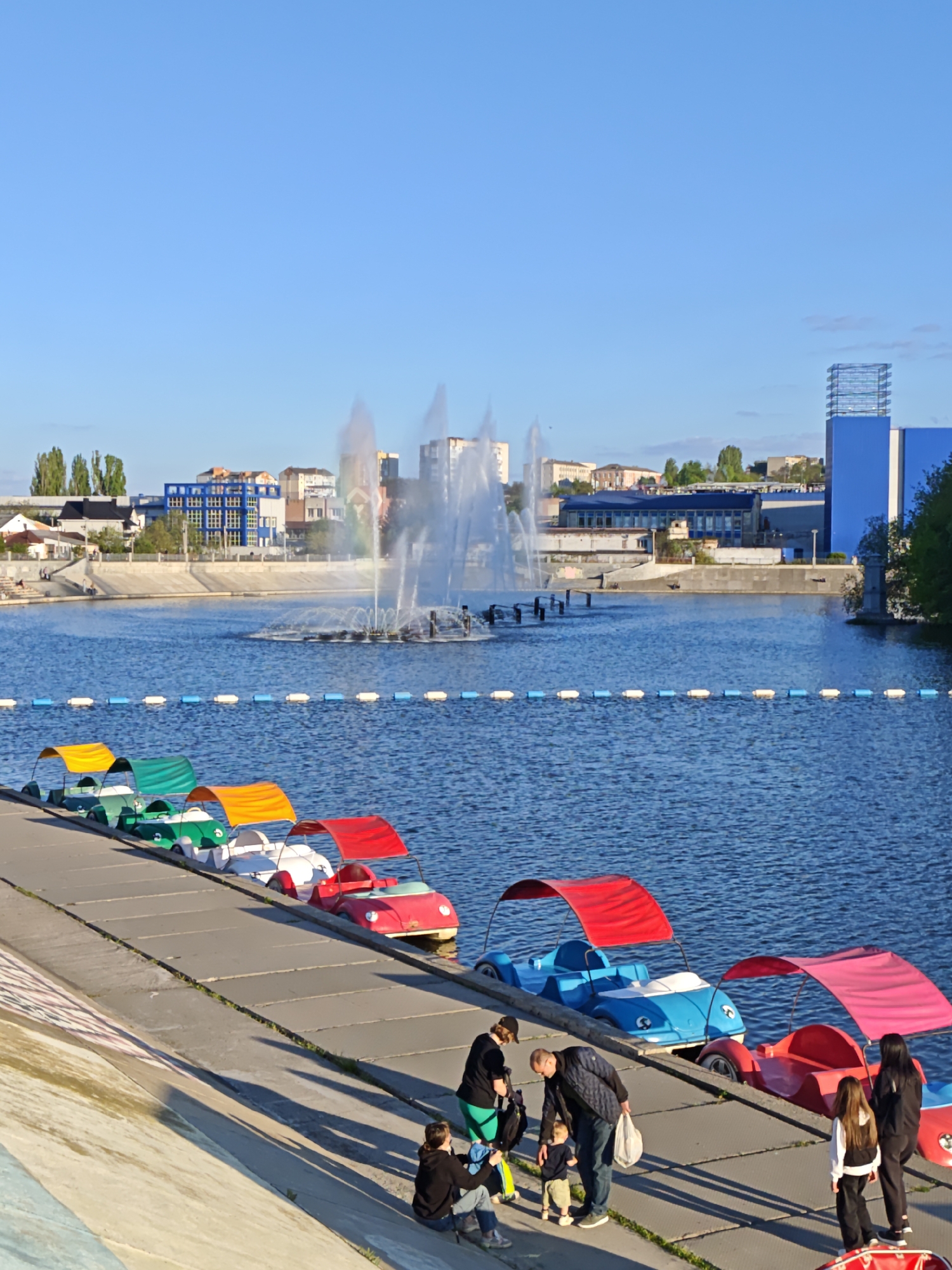 The image depicts a waterfront scene in Vinnytsia, Ukraine, featuring a body of water with a fountain in the center. The fountain is spraying water upwards, creating a rainbow effect. A line of colorful paddle boats is docked along a concrete embankment. A group of people, including adults and children, are gathered near the paddle boats. The background includes buildings and trees. The sky is clear and blue. The overall scene suggests a leisure or recreational area within the city.