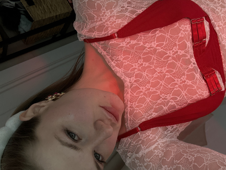 A woman in a white lace bodysuit with red straps poses. She wears an ornate gold earring and a white headband. The image is dimly lit, with a red cast on her face and neck. Part of a woven basket is visible in the background. The photo is taken at an angle, making the woman appear sideways.